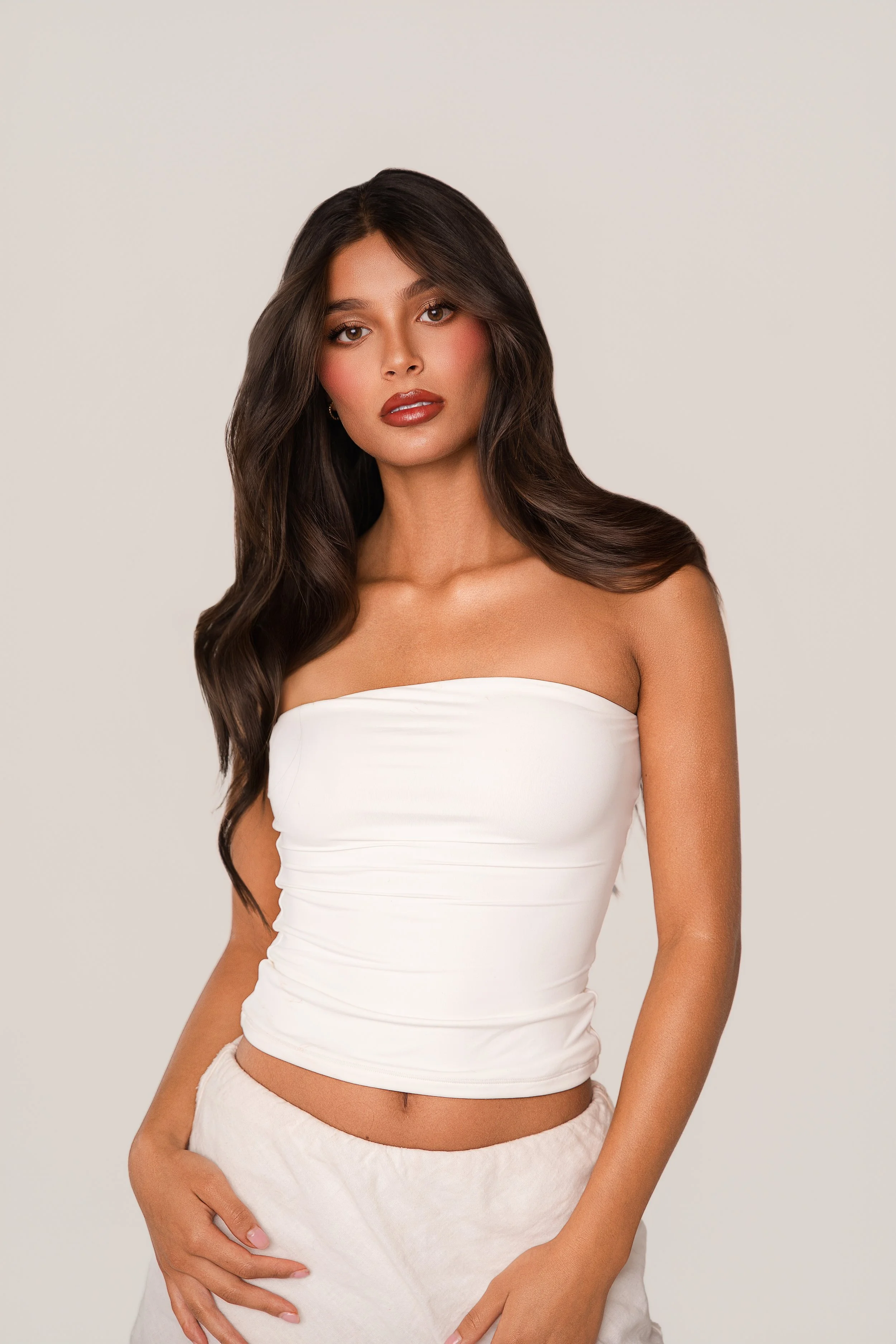A young woman with long, wavy dark hair, wearing a strapless white top and light-colored pants, standing against a plain background.