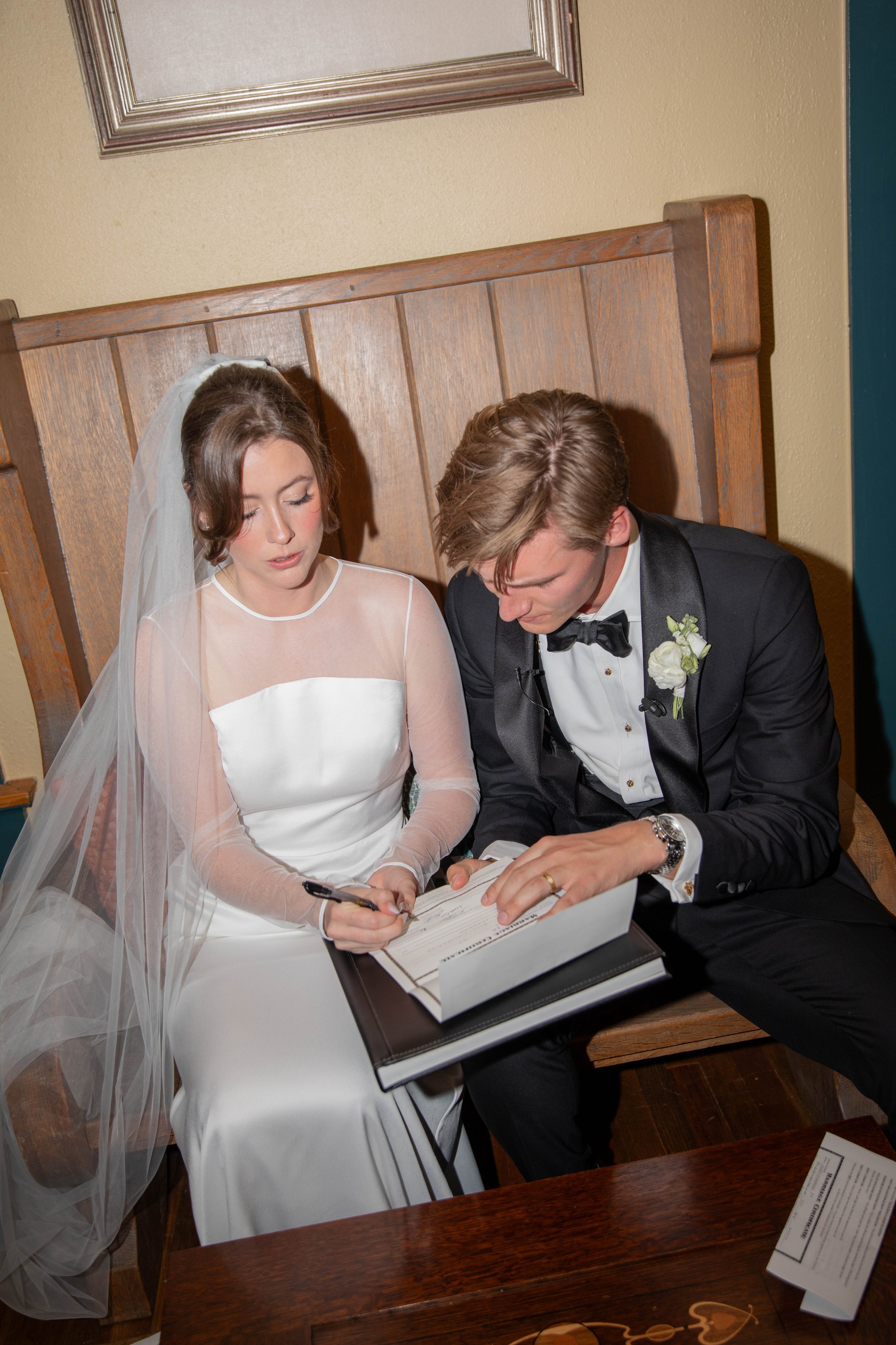 A bride and groom sitting together, signing a marriage document at their wedding ceremony, with the bride wearing a white wedding dress and veil, and the groom in a black tuxedo with a boutonniere.