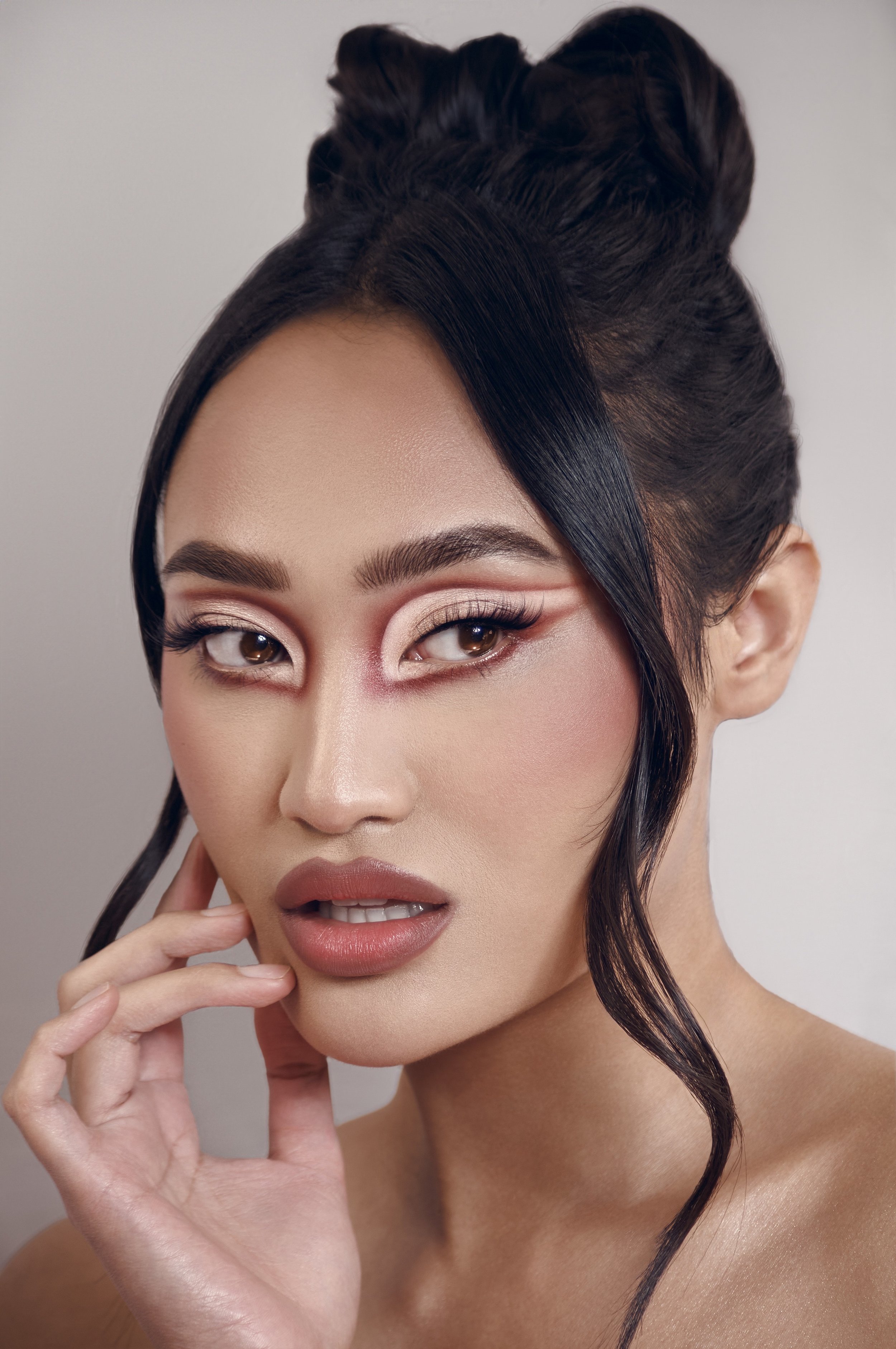 Close-up of a woman with styled hair, makeup, and a confident expression, touching her face lightly with her hand.