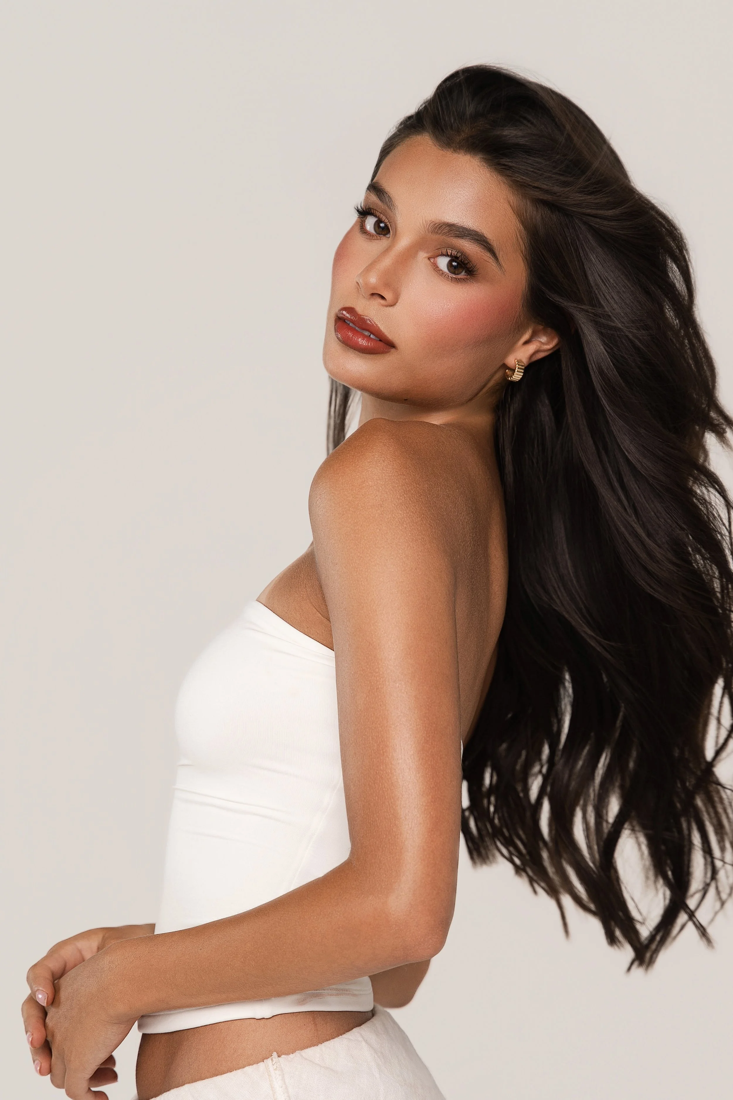 A young woman with long, wavy dark hair and light brown skin, wearing a white tube top and light-colored pants, posing against a plain light background.