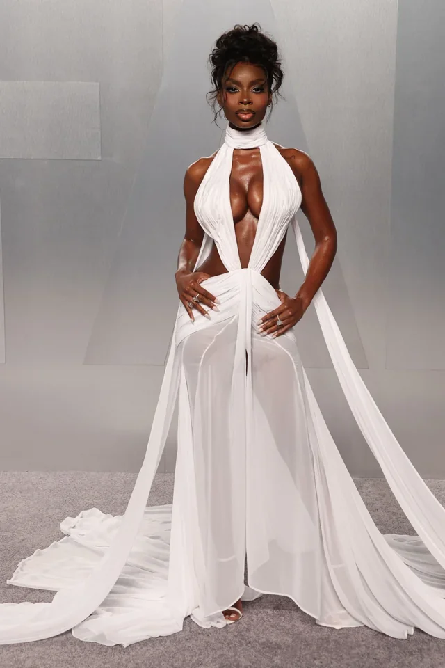 Olandria with curly hair in an elaborate white gown with cutouts and long flowing fabric, posing confidently against a plain gray backdrop at Vanity Fair