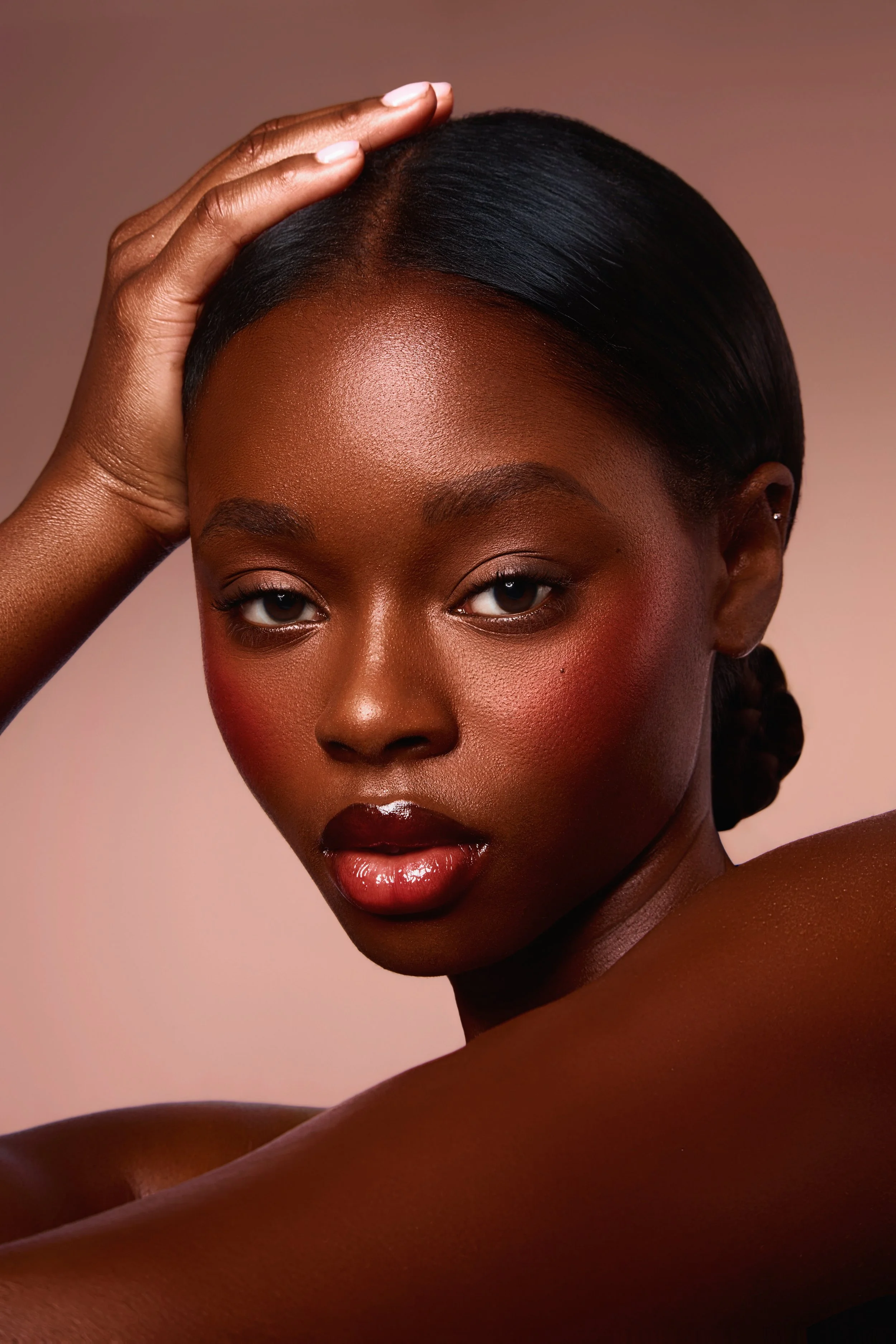 A close-up portrait of a Black woman with smooth skin and dark hair styled in a sleek bun. She has subtle eye makeup, glossy lips, and a confident expression, with one hand resting on her head.