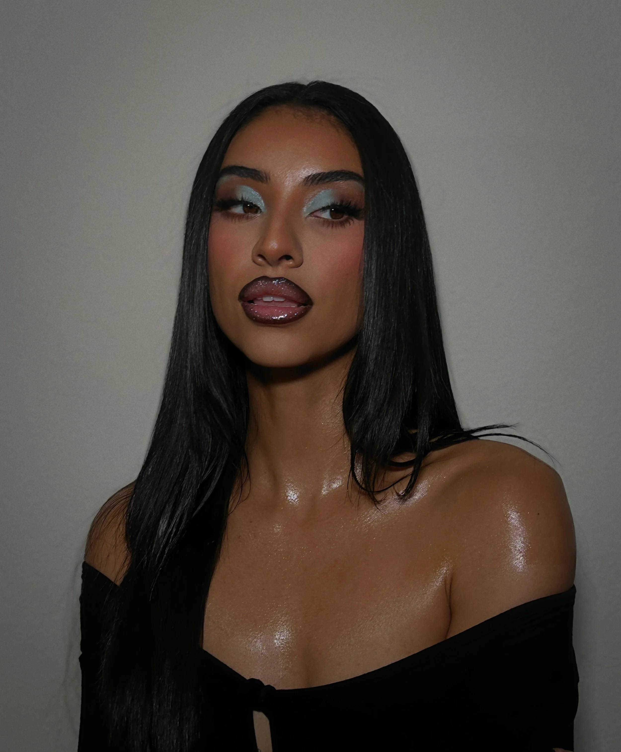 A woman with long black hair wearing makeup, glossy dark lipstick, light blue eyeshadow, and an off-shoulder black top, posing against a plain light-colored wall.