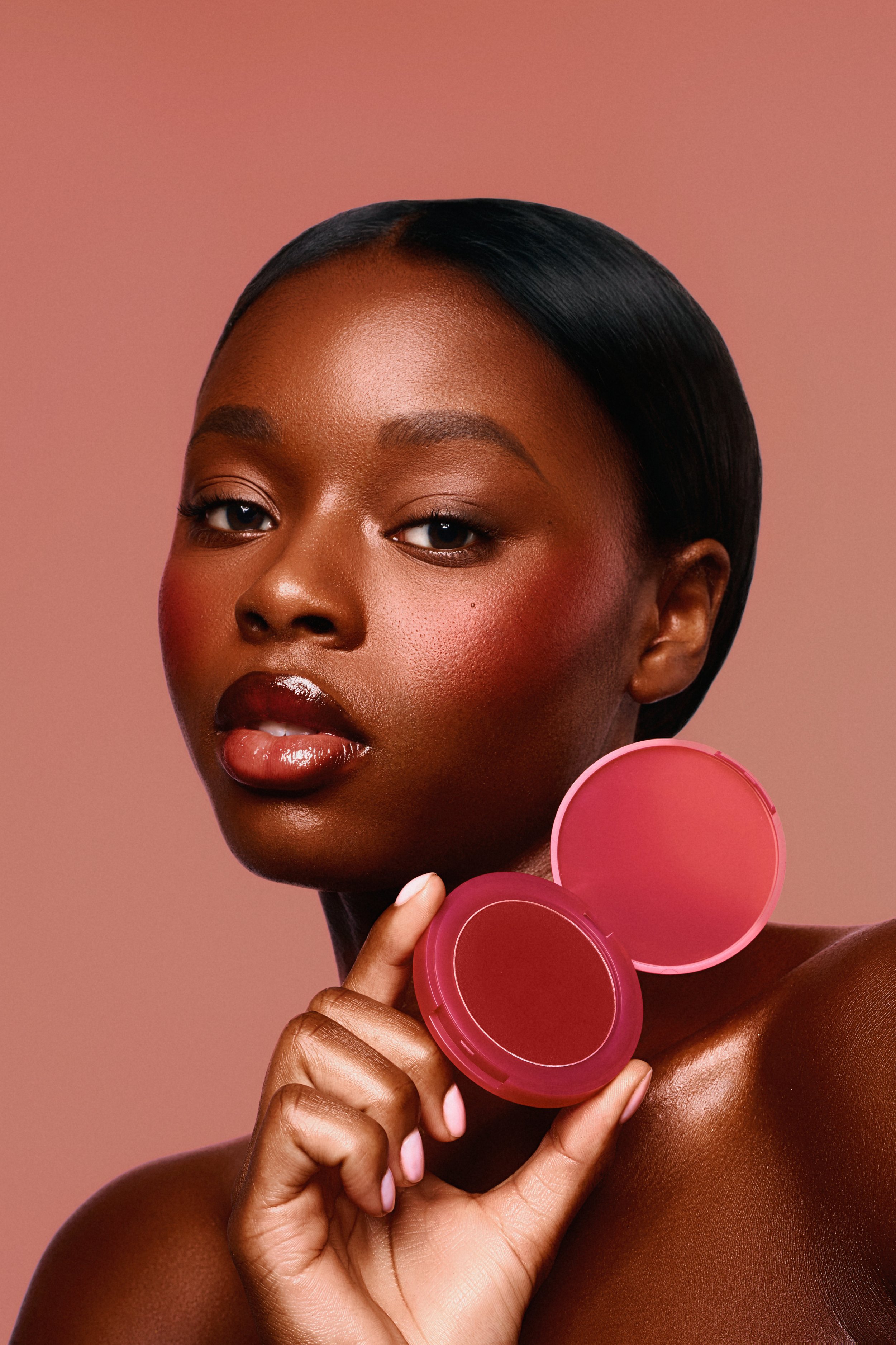 A woman with dark skin and short, sleek black hair holds a pink makeup compact near her face, with a pink background.
