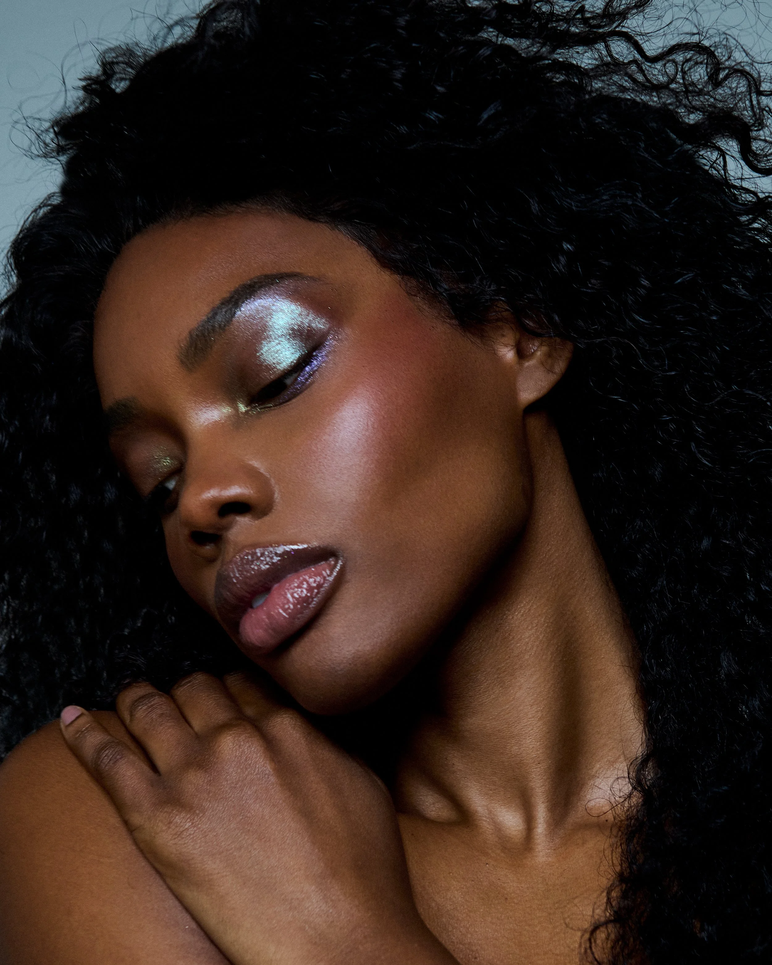 Close-up of a woman with curly black hair, wearing shiny makeup, including metallic eyeshadow, glossy lips, and highlighted cheekbones, touching her shoulder with her hand.