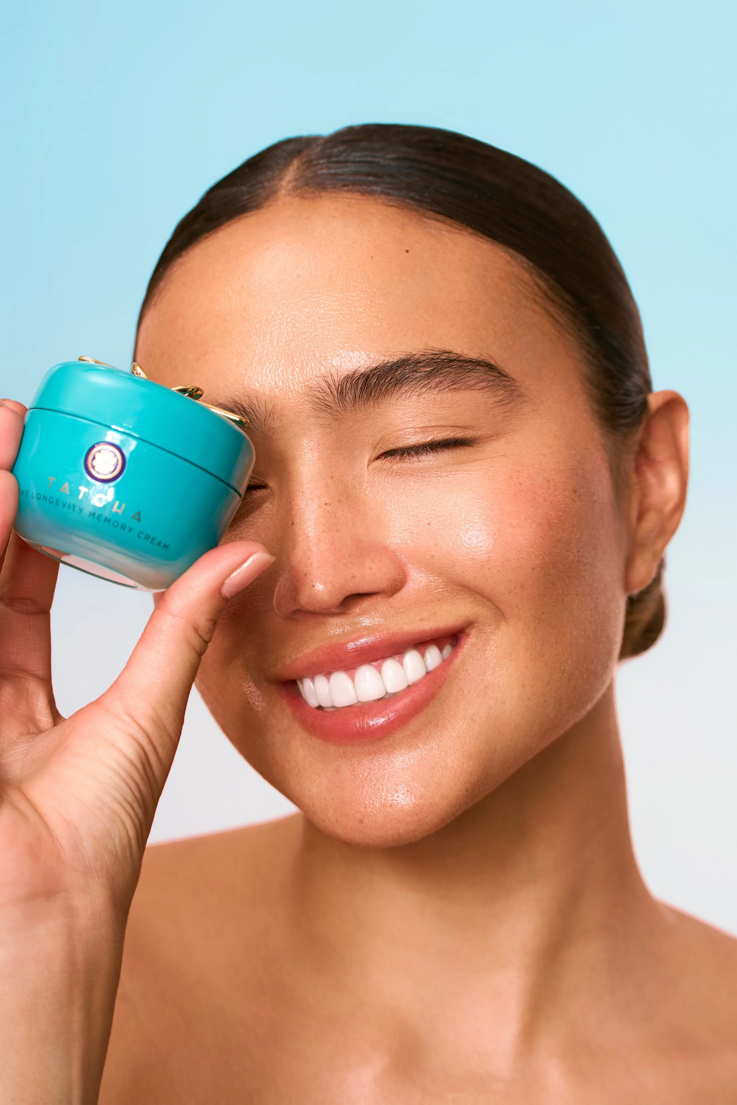 A woman with clear skin and a bright smile holding a small, blue jar of skincare cream near her eye, against a pastel background.
