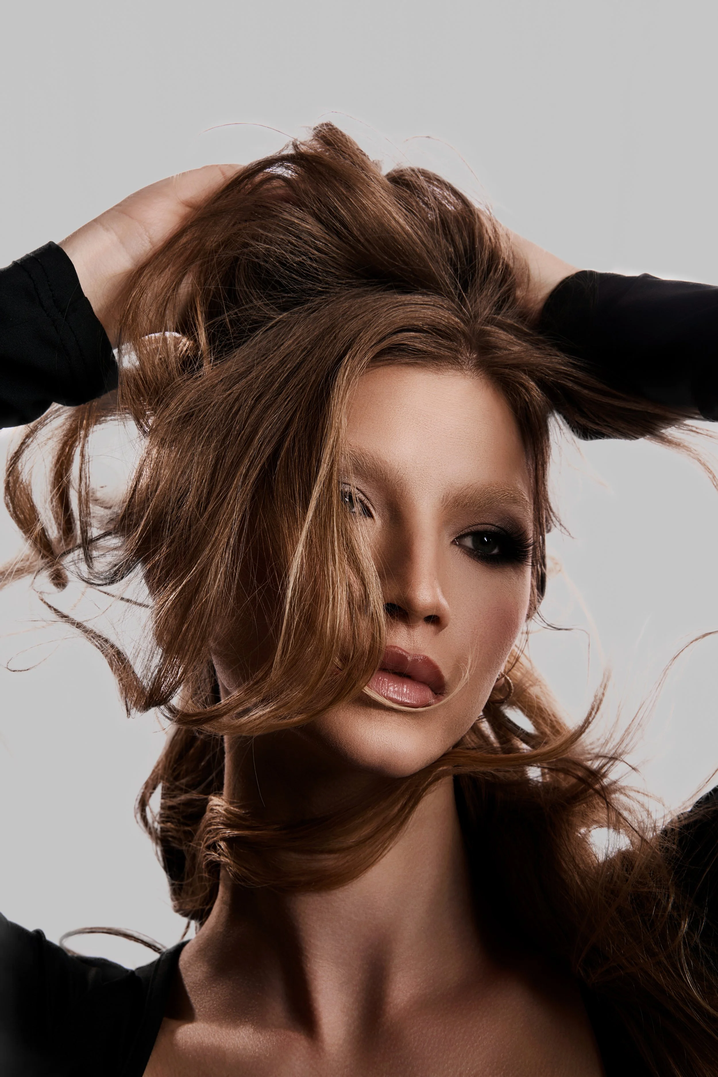 A woman with straight, light brown hair, styled with waves, partially covering one eye, posing with her hands in her hair, against a plain background.