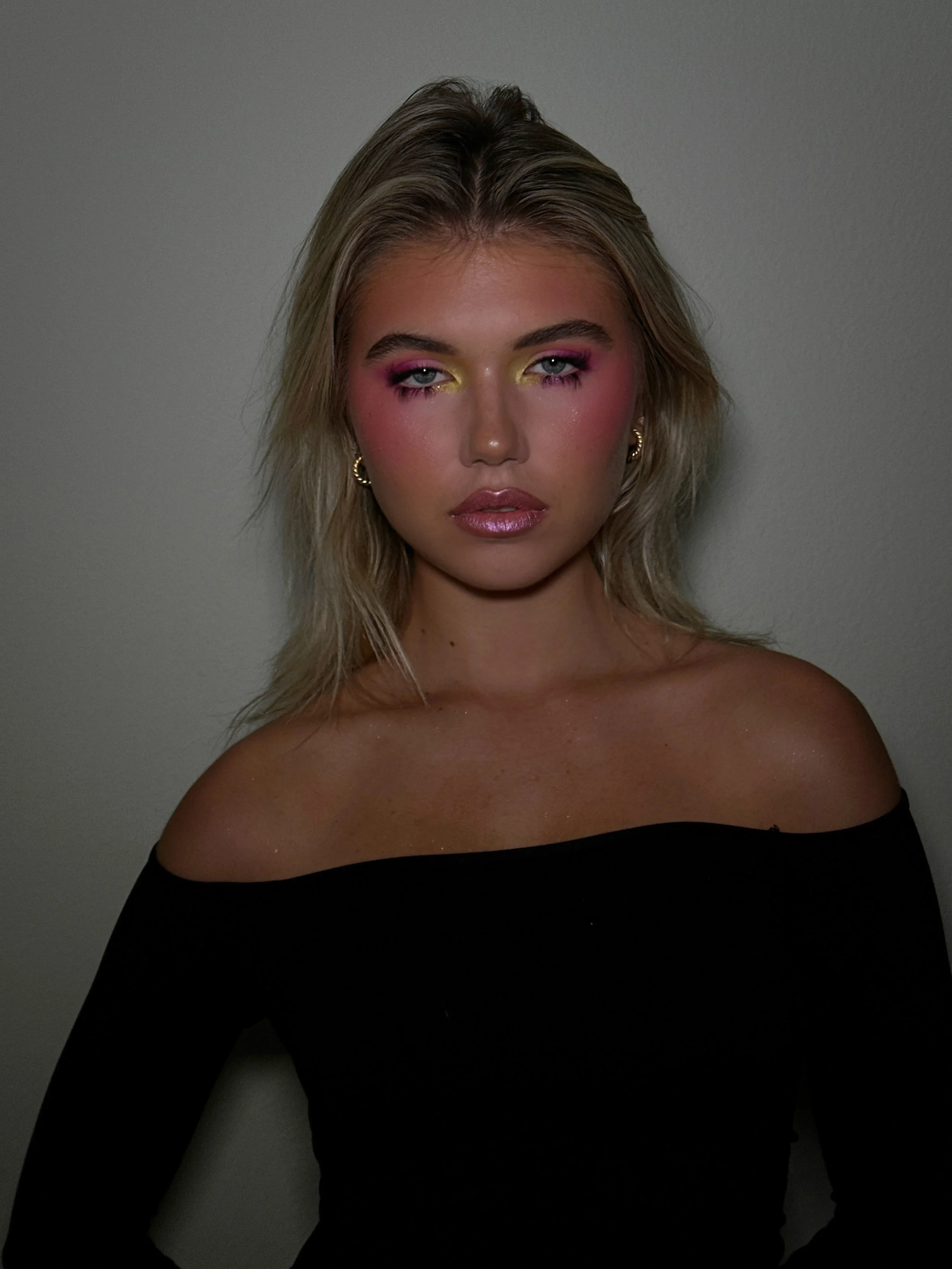 A woman with blonde hair and colorful eye makeup, wearing gold hoop earrings and an off-the-shoulder black top, standing against a plain wall.