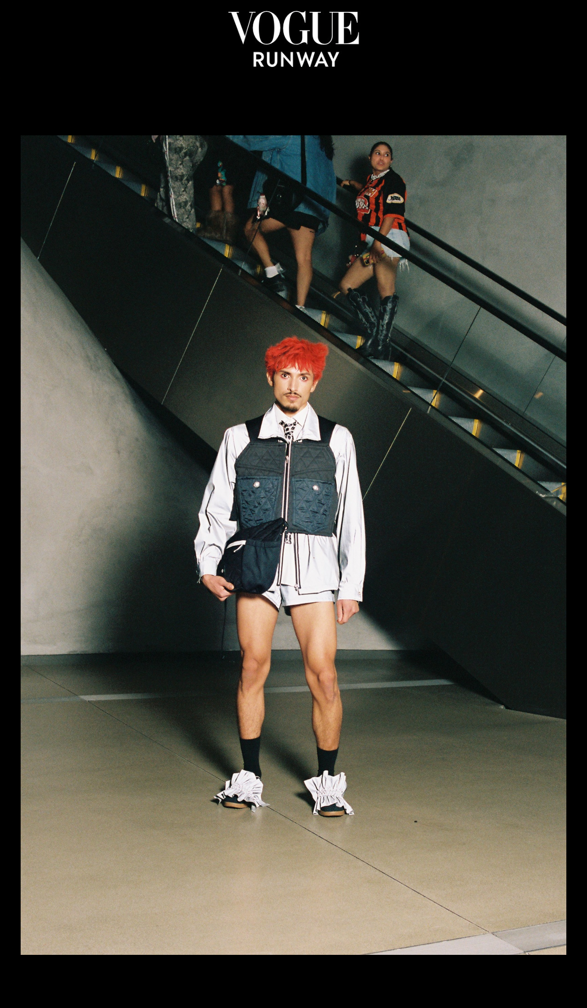 A man with red hair and a goatee wearing a white jacket, black shorts, and ruffled sneakers. He is standing in an indoor space with a modern concrete wall and an escalator in the background. A woman in a racing-themed outfit is on the escalator behin