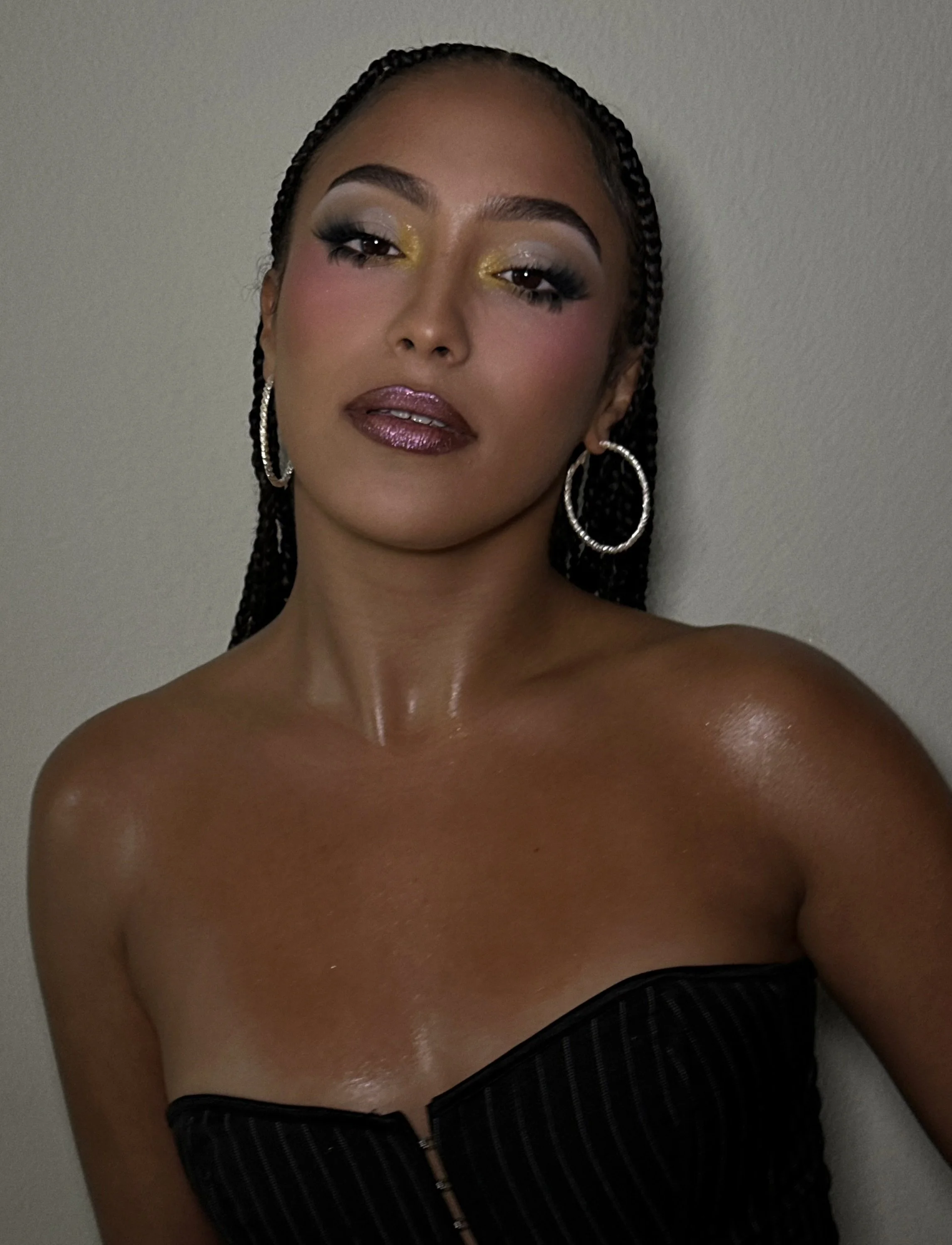 A woman with makeup and long braided hair wearing hoop earrings and a black strapless top standing against a plain wall.