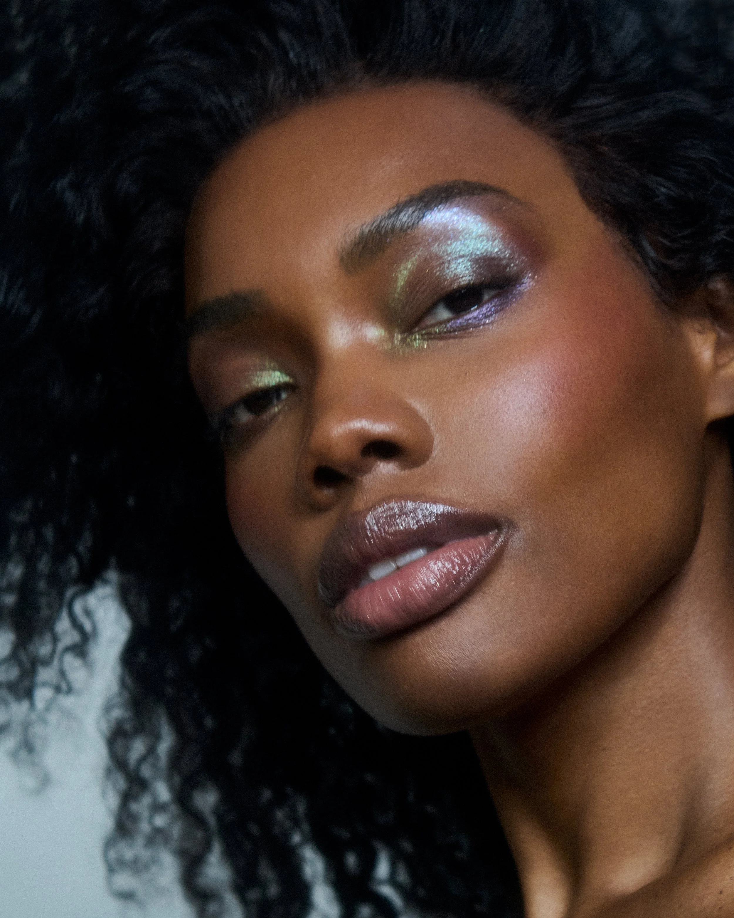 Close-up of a woman with dark skin and curly black hair, wearing shimmery eyeshadow, glossy lipstick, and glowing skin.