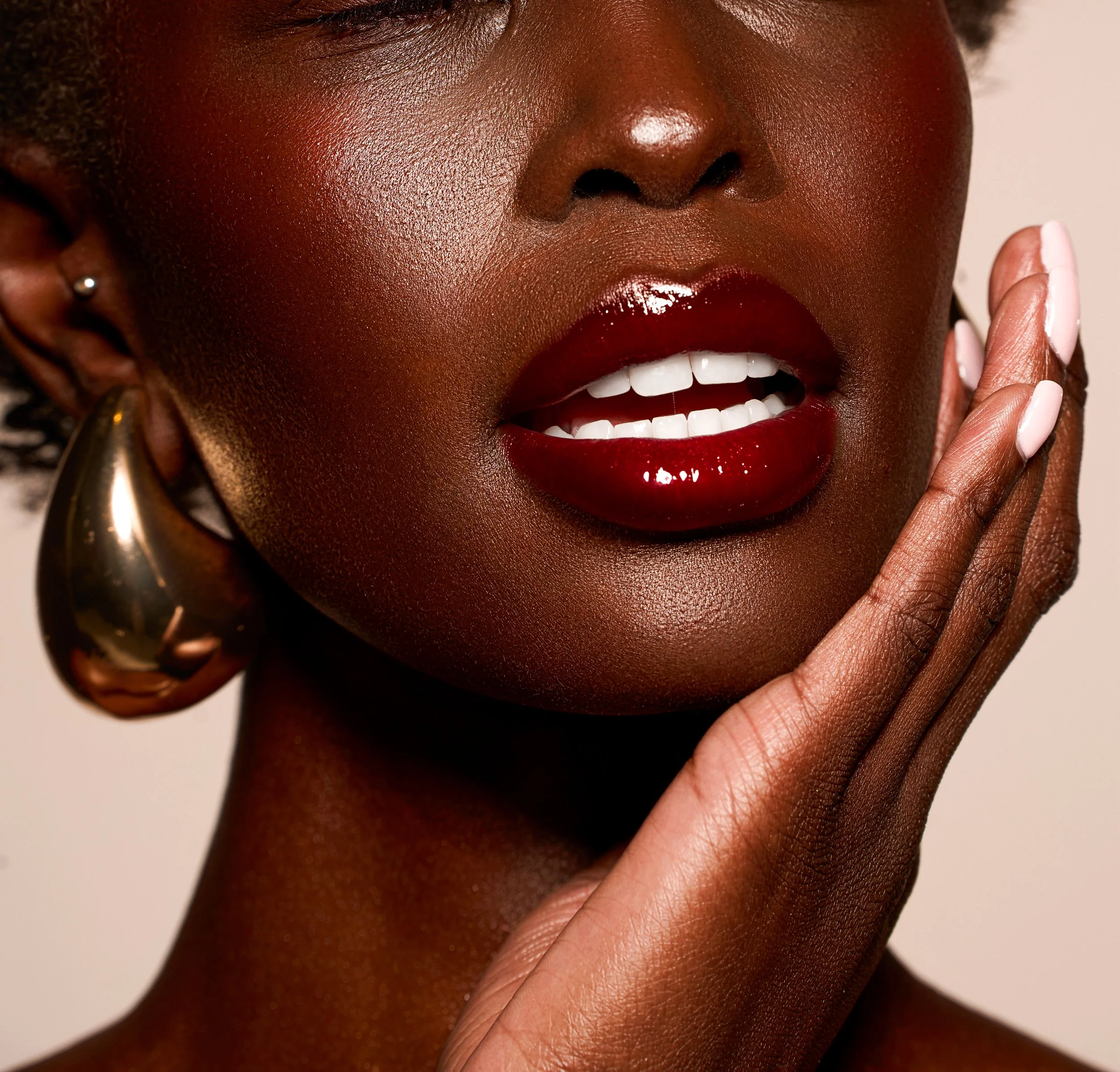 Close-up of a woman with dark skin wearing large gold hoop earrings, dark red lipstick, and glossy makeup. She has short hair and rests her hand on her face, showing painted nails.
