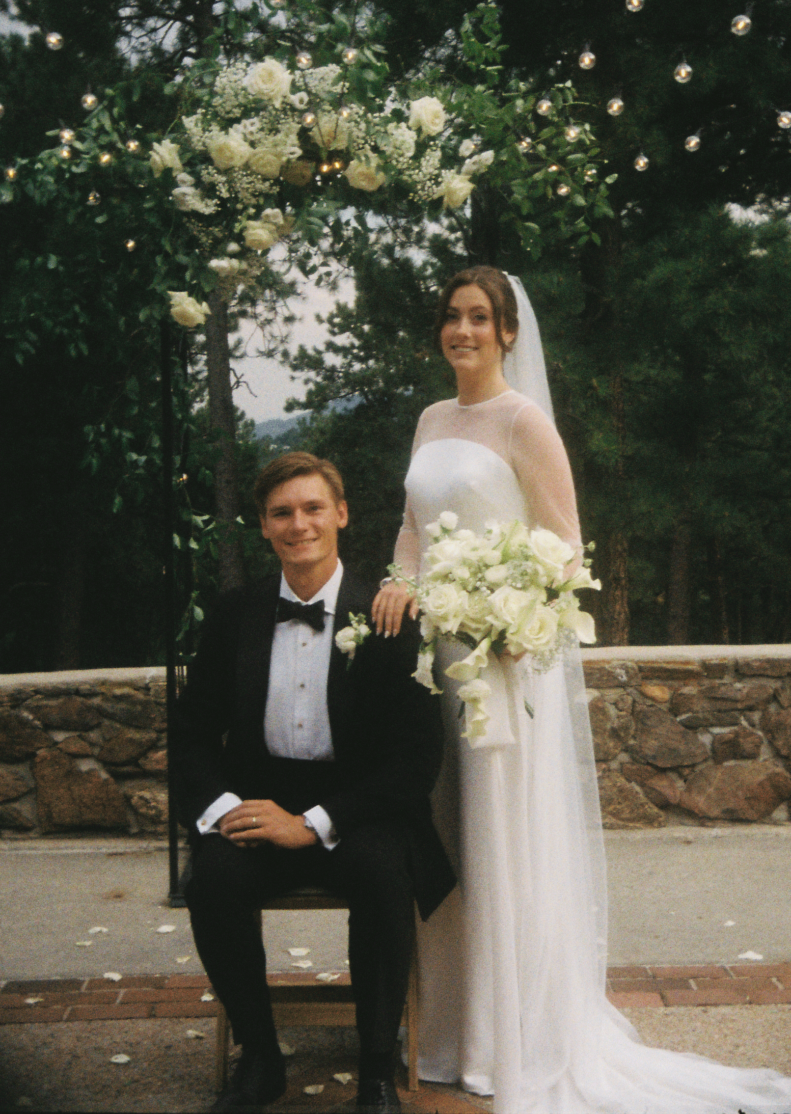 A woman in a white wedding dress and veil stands behind a seated man in a black tuxedo, both smiling, in front of a decorated outdoor wedding arch with flowers and string lights.