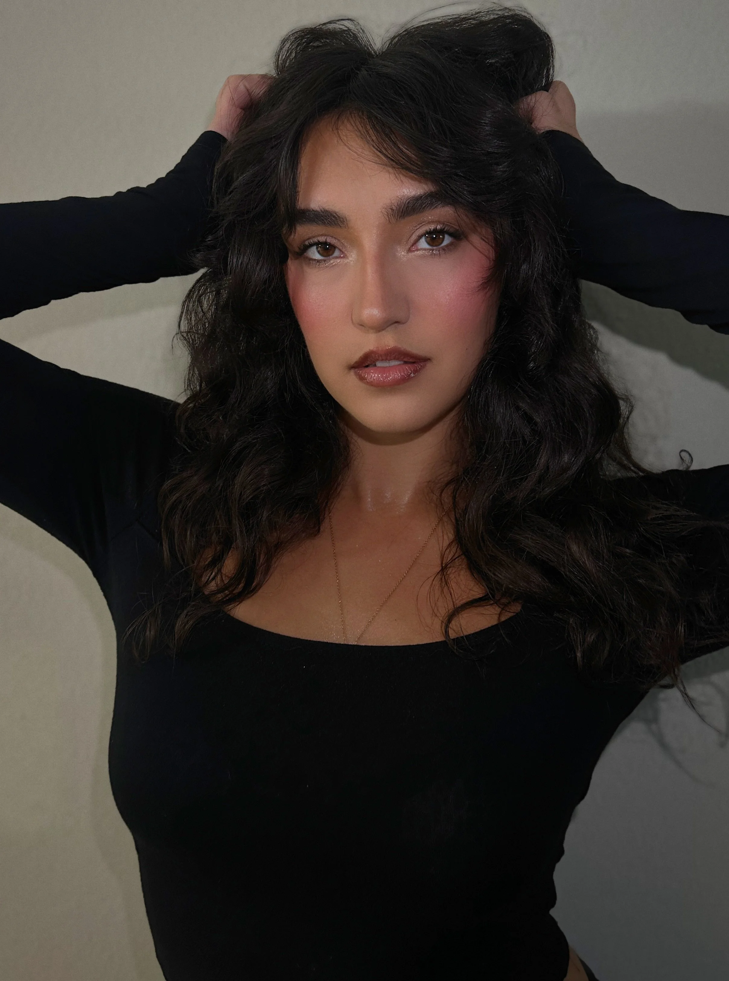 A woman with long, wavy dark hair and light skin, wearing a black top, with her hands behind her head, looking at the camera with a confident expression.