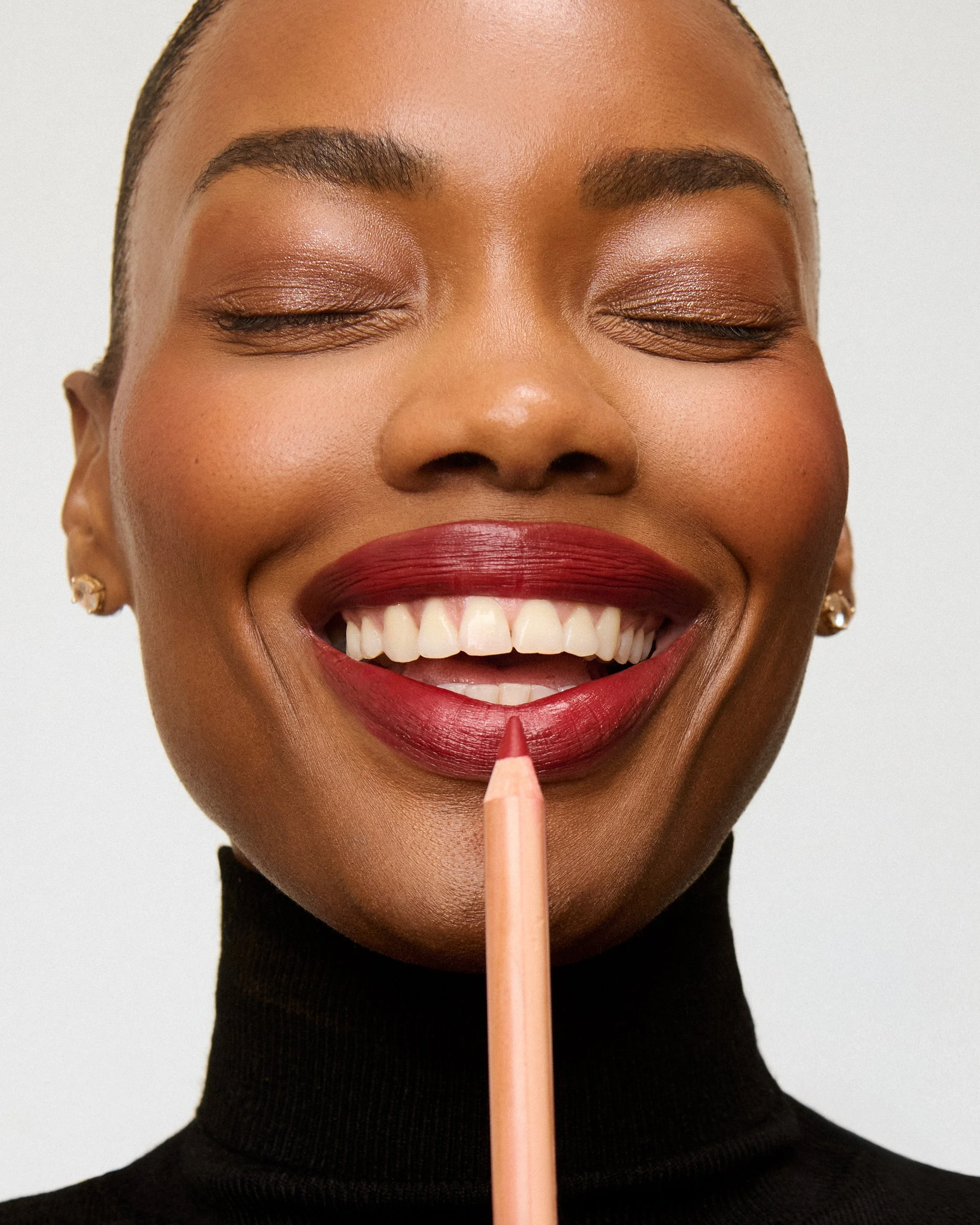Close-up of a woman with her eyes closed, smiling, holding a lipstick pencil against her lips, wearing a black turtleneck and earrings.