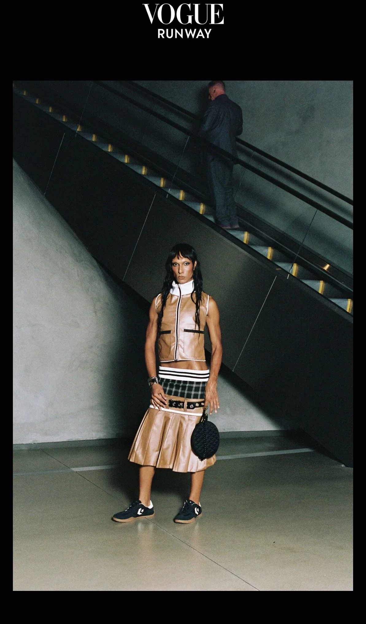 Fashion model standing indoors in front of a concrete wall with a staircase above, wearing a beige and tan vest, a plaid skirt, black athletic shoes, and carrying a black round bag, with a man walking up the staircase in the background.