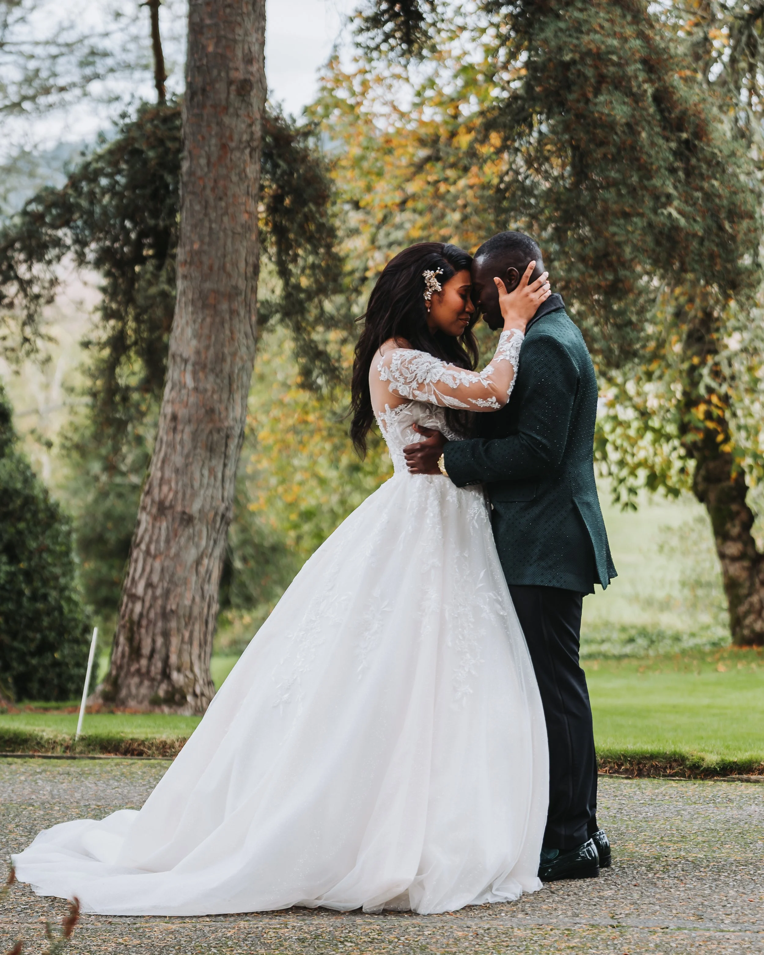 A bride in a white wedding gown and a groom in a dark suit share an intimate moment outdoors among trees and greenery, with their foreheads touching and eyes closed.