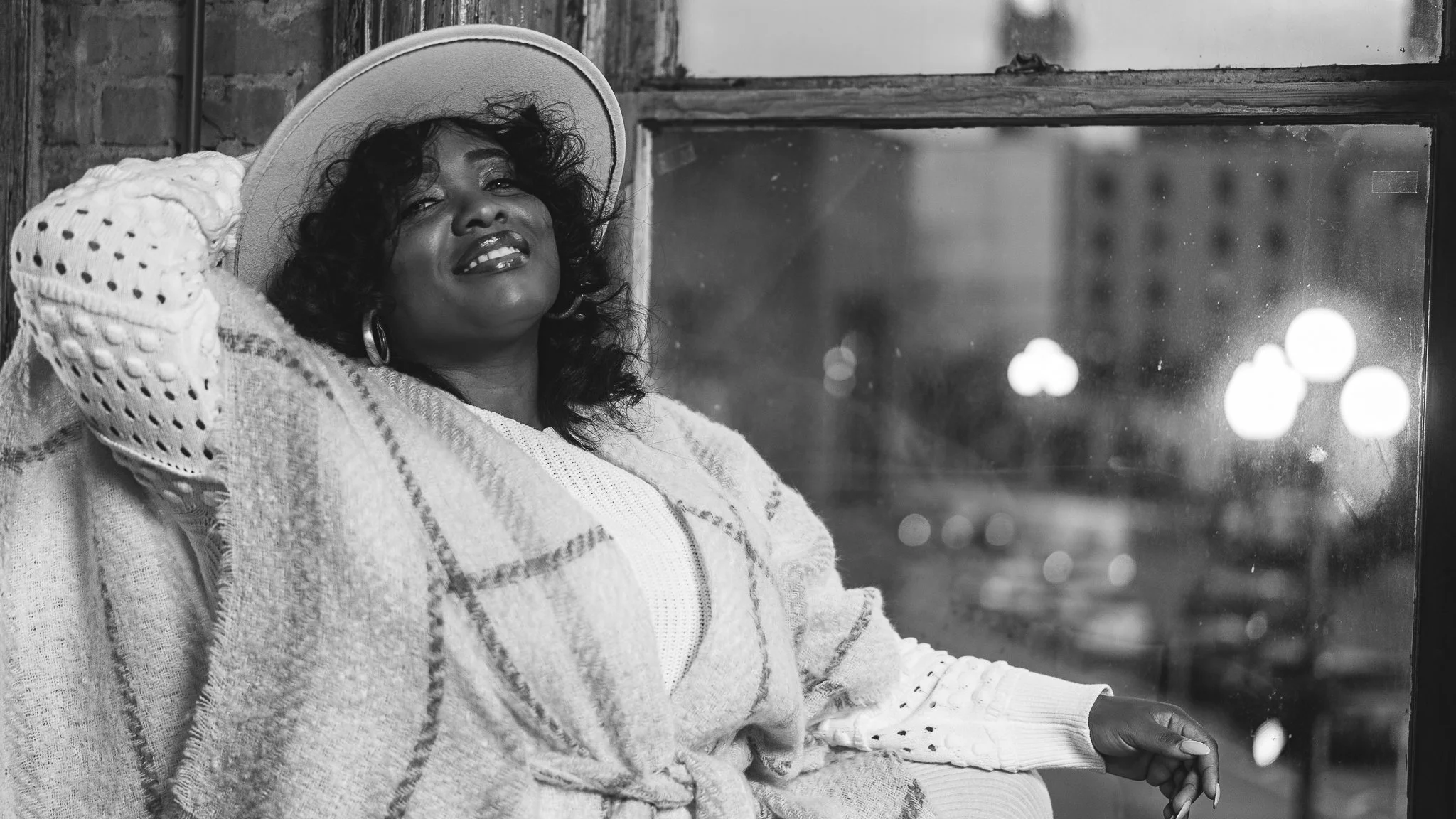 A woman with curly hair wearing a wide-brimmed hat, hoop earrings, a cozy sweater, and a patterned shawl, sitting by a large window in an urban setting at night with city lights outside.