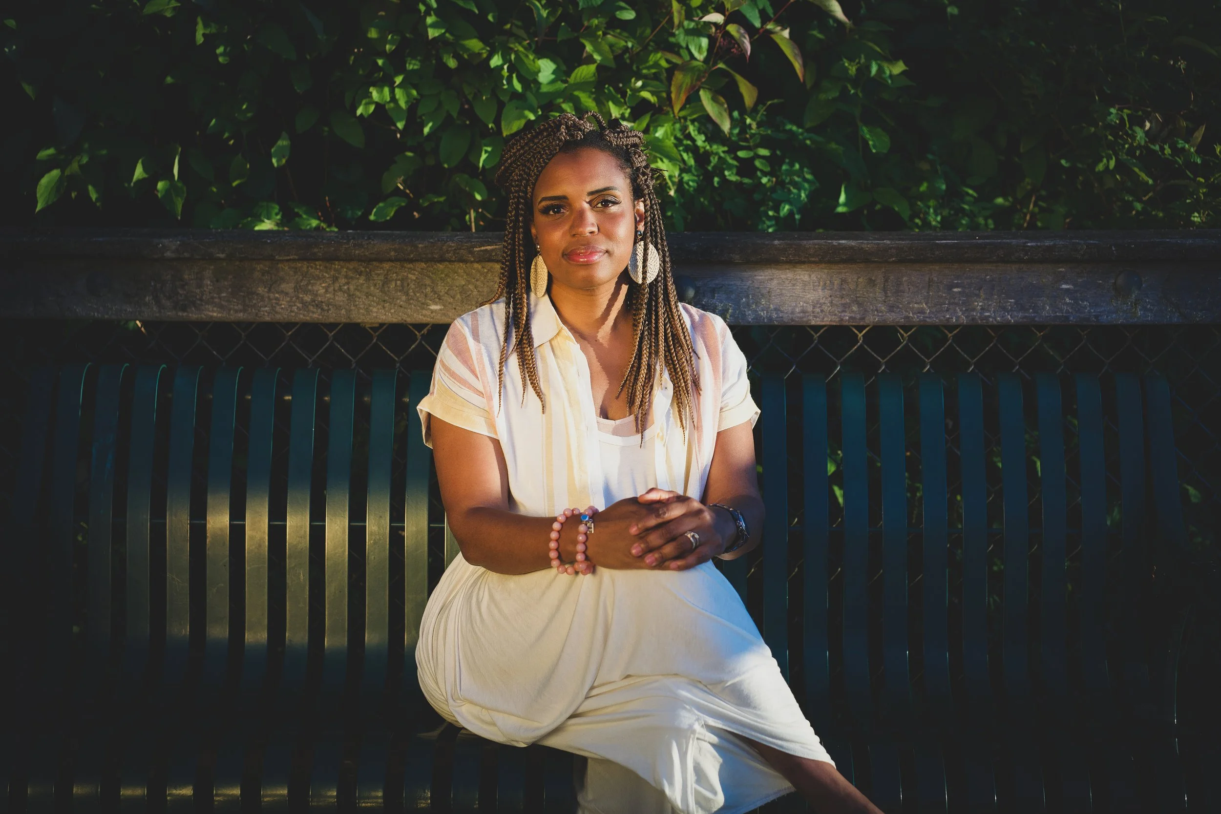 A woman with braided hair sits on a park bench, wearing a light-colored dress and large earrings. She is outdoors, with lush green foliage behind her, and is looking directly at the camera with her hands clasped in her lap.