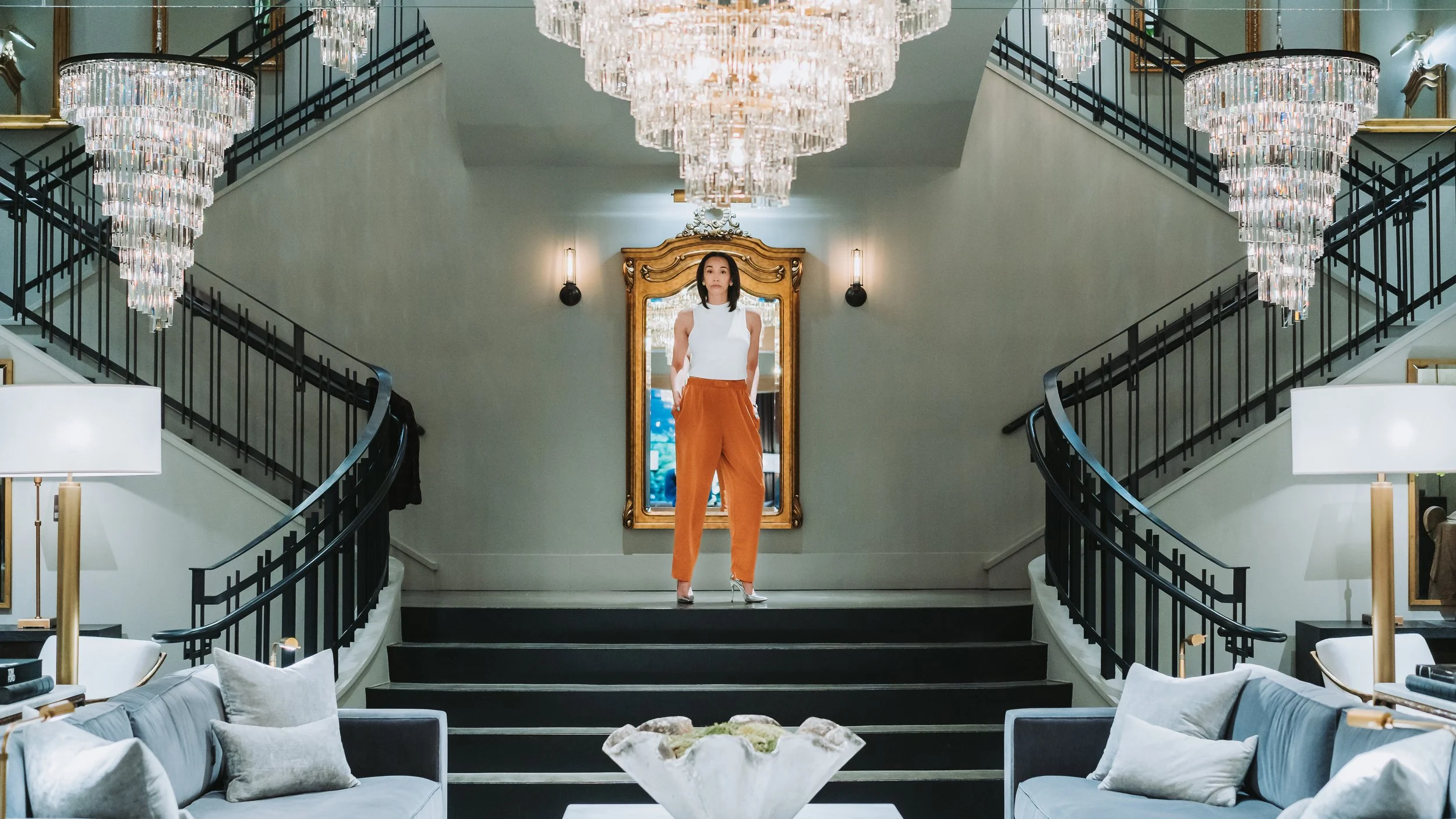 A woman standing on a staircase in an elegant, chandelier-lit room with a mirror behind her.