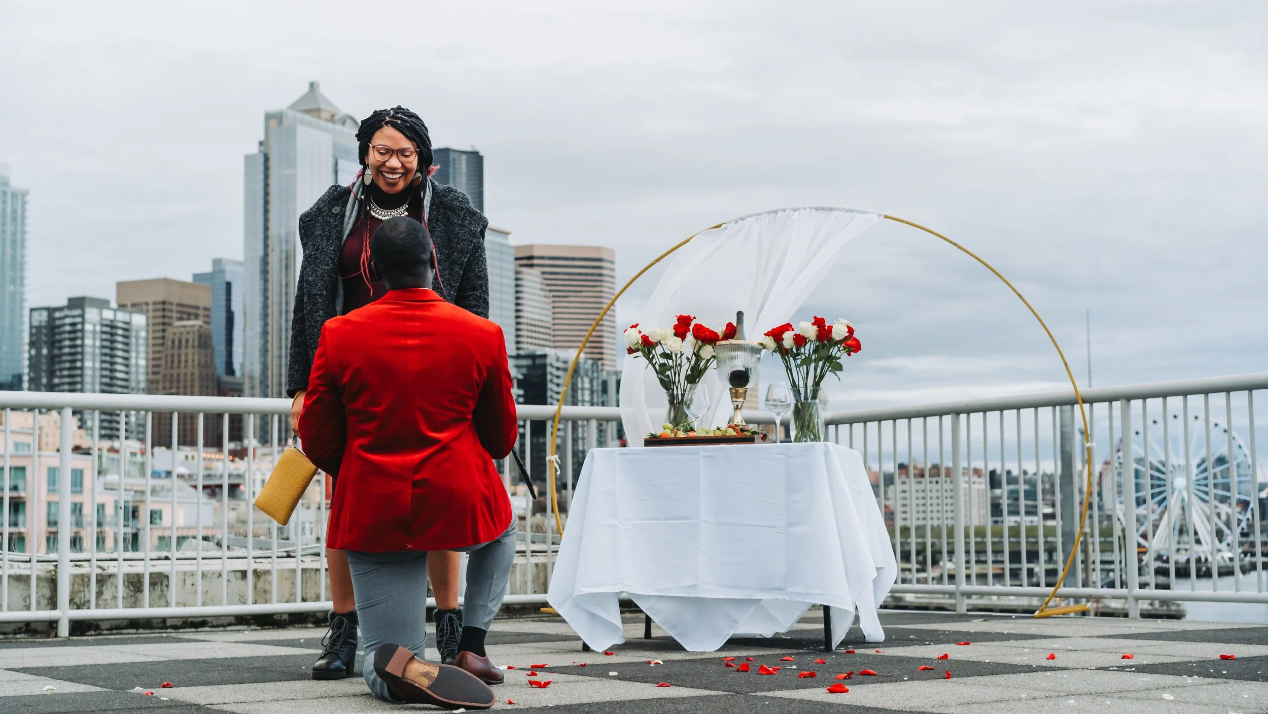 A woman is proposing marriage to a man on a rooftop with a city skyline in the background. The man is kneeling, and there is a decorated table with flowers, wine, and dessert nearby.