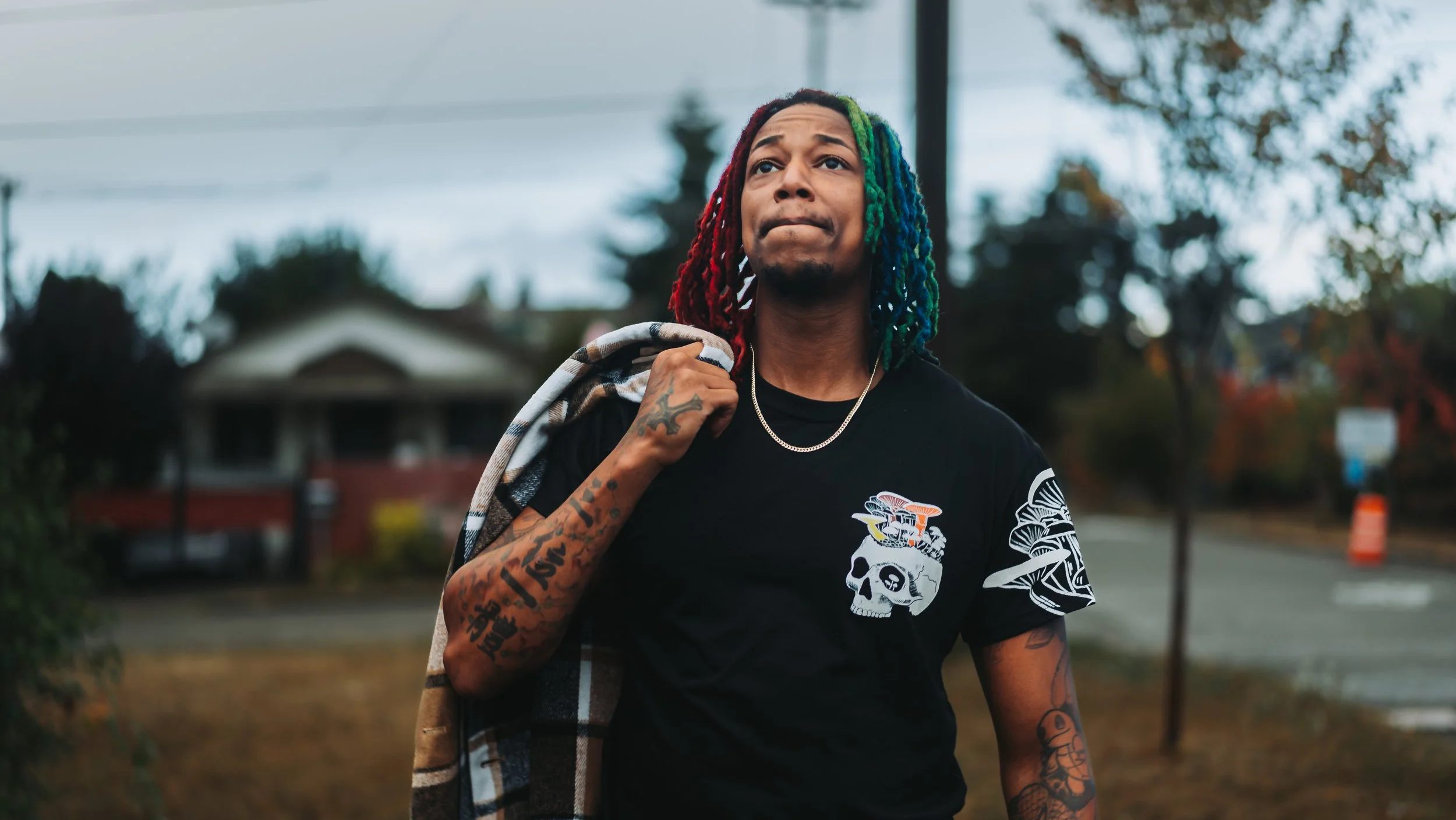 A young man with multi-colored dreadlocks, wearing a black T-shirt with a skull graphic, holding a plaid jacket over his shoulder, walking outdoors on a cloudy day.