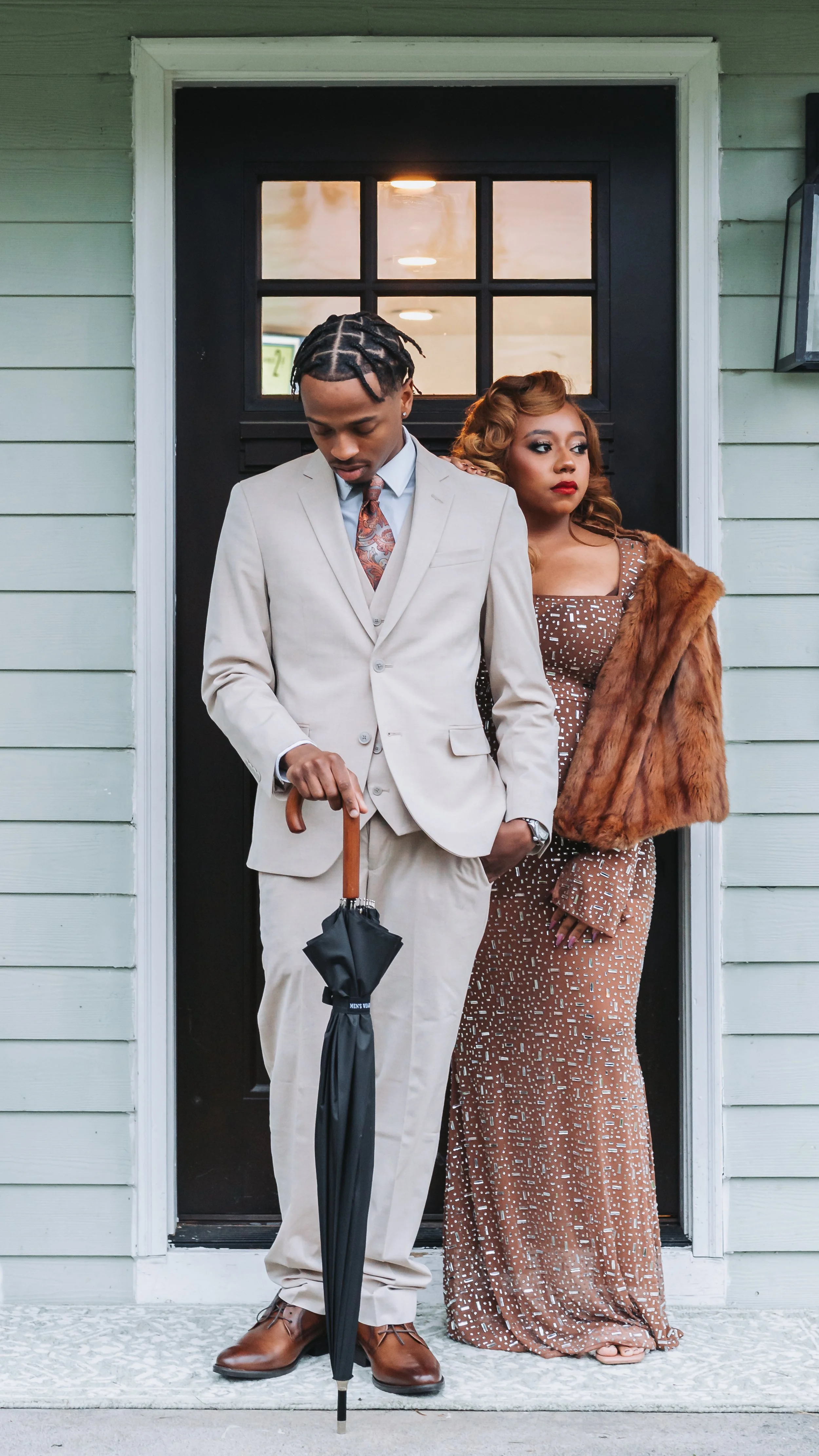 A man and woman standing in front of a house door. The man is looking down, holding a black umbrella, wearing a beige suit with a patterned tie. The woman leans slightly against the door, wearing a brown polka dot dress and a brown fur coat.