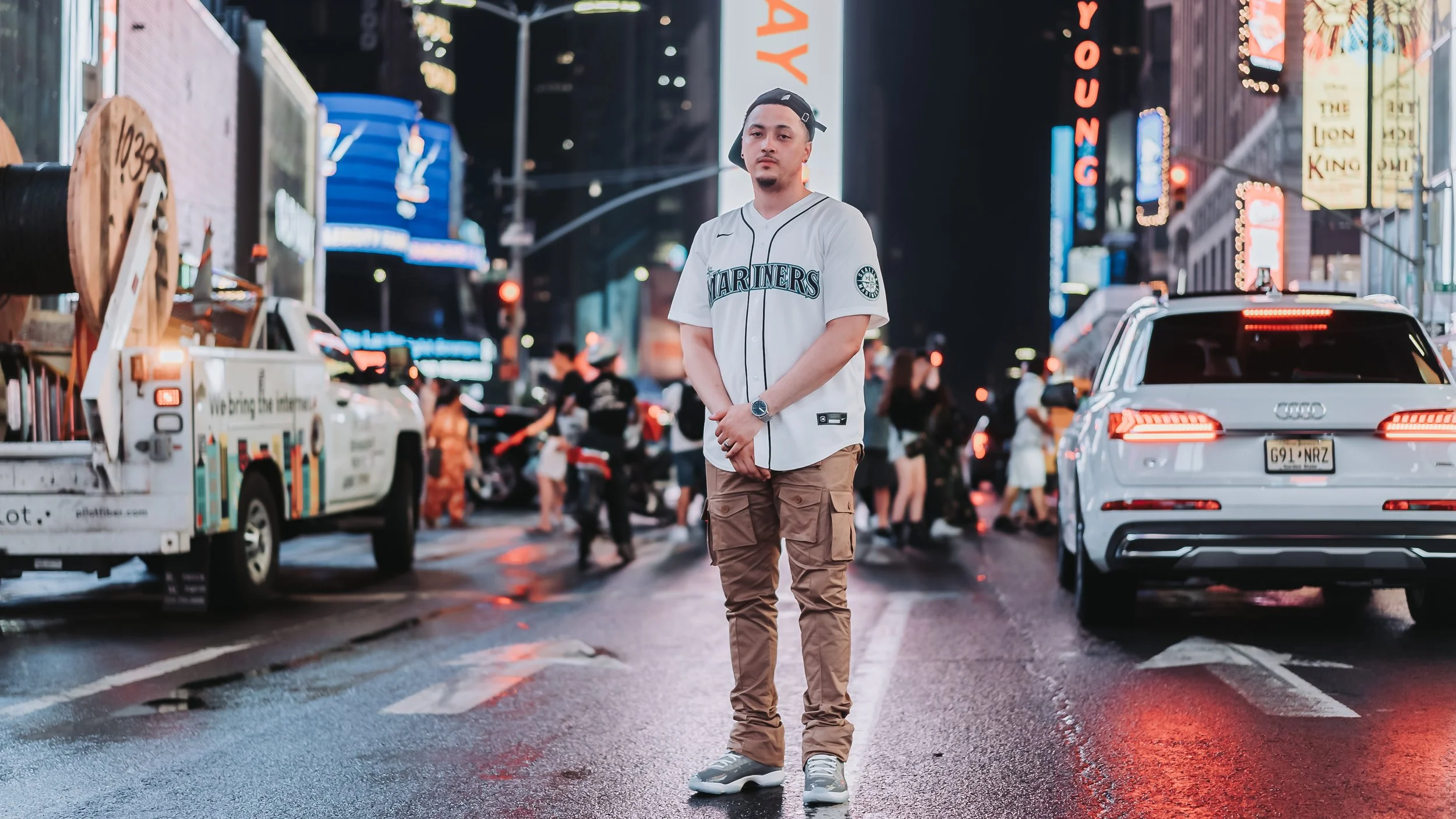 A man standing in the middle of a busy city street at night, wearing a Seattle Mariners baseball jersey, tan cargo pants, and sneakers, with a watch on his left wrist, surrounded by cars, trucks, and pedestrians, illuminated by neon signs and city li