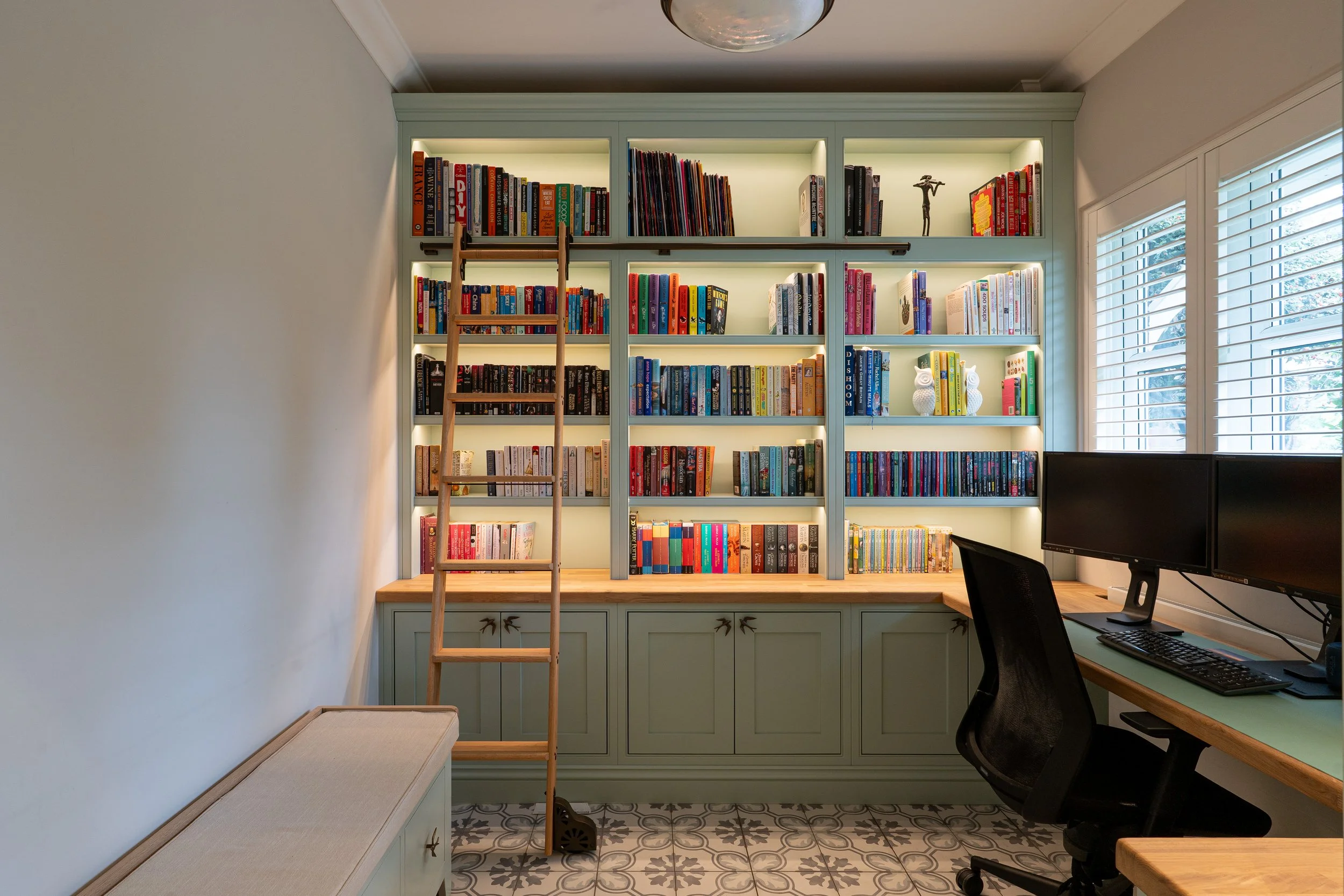 A room with a light green bookshelf filled with colorful books, a wooden ladder to reach higher shelves, a window with white blinds, a desk with two computer monitors and a black chair, a bench with a cushion, and patterned floor tiles.