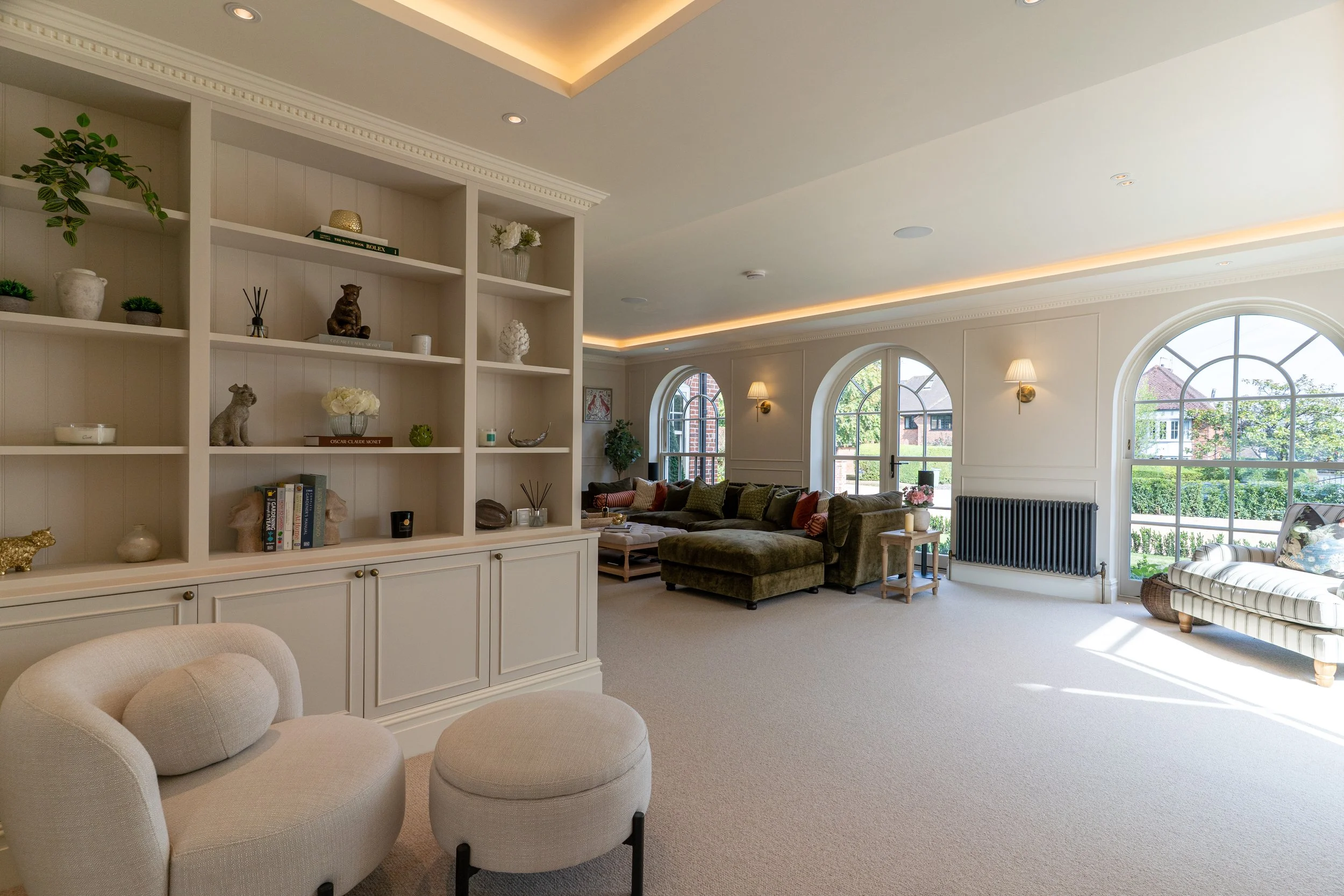 A spacious living room with large arched windows letting in natural light, a beige carpet, and a variety of furniture including a rounded cream-colored chair, a green velvet sectional sofa, and a white striped armchair. Decor includes a bookshelf wit
