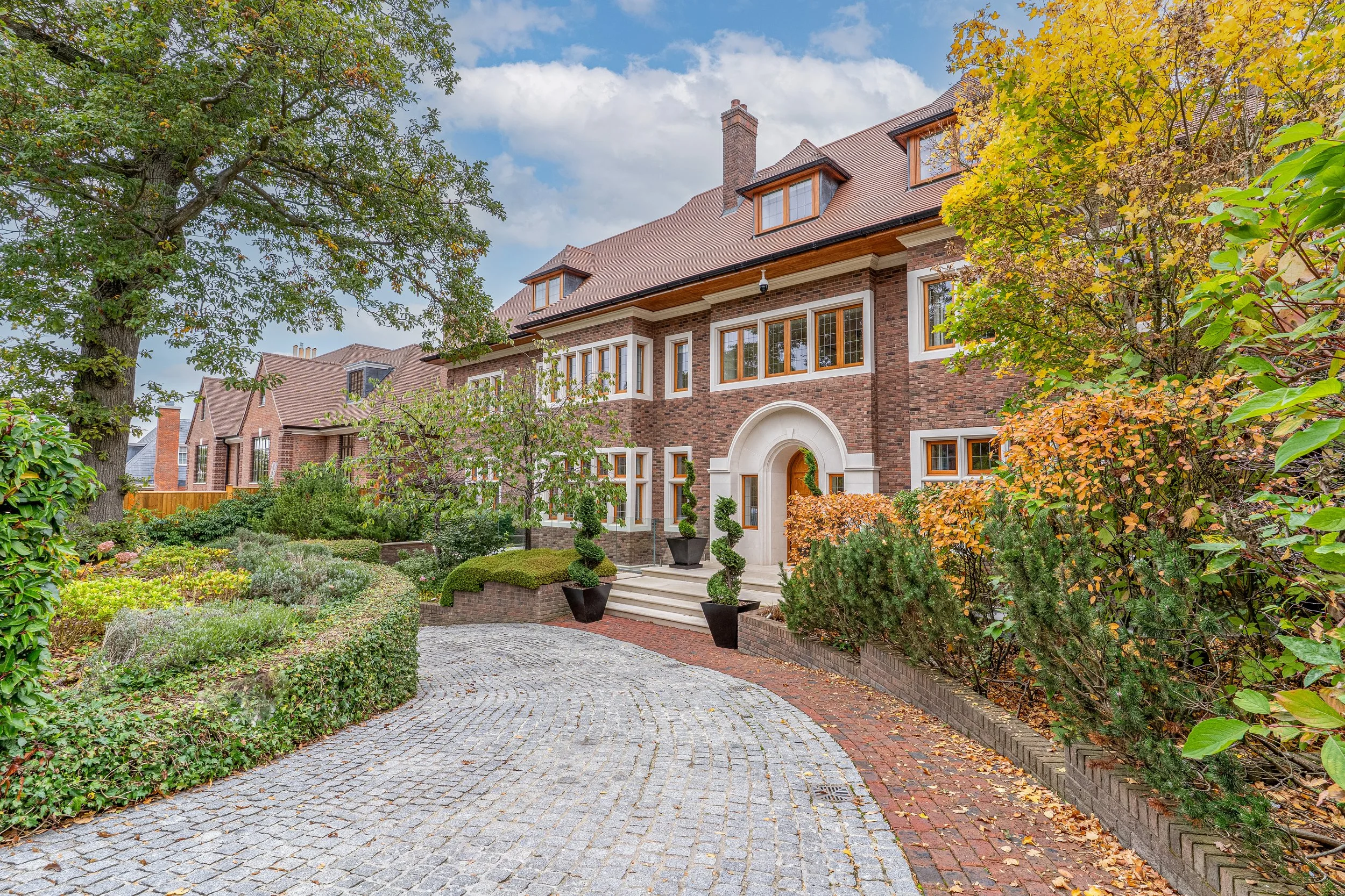 A large brick house with multiple gabled roofs surrounded by a lush, well-maintained garden with various trees and shrubs. A curved cobblestone driveway leads to the entrance, which has white stone steps and potted plants.