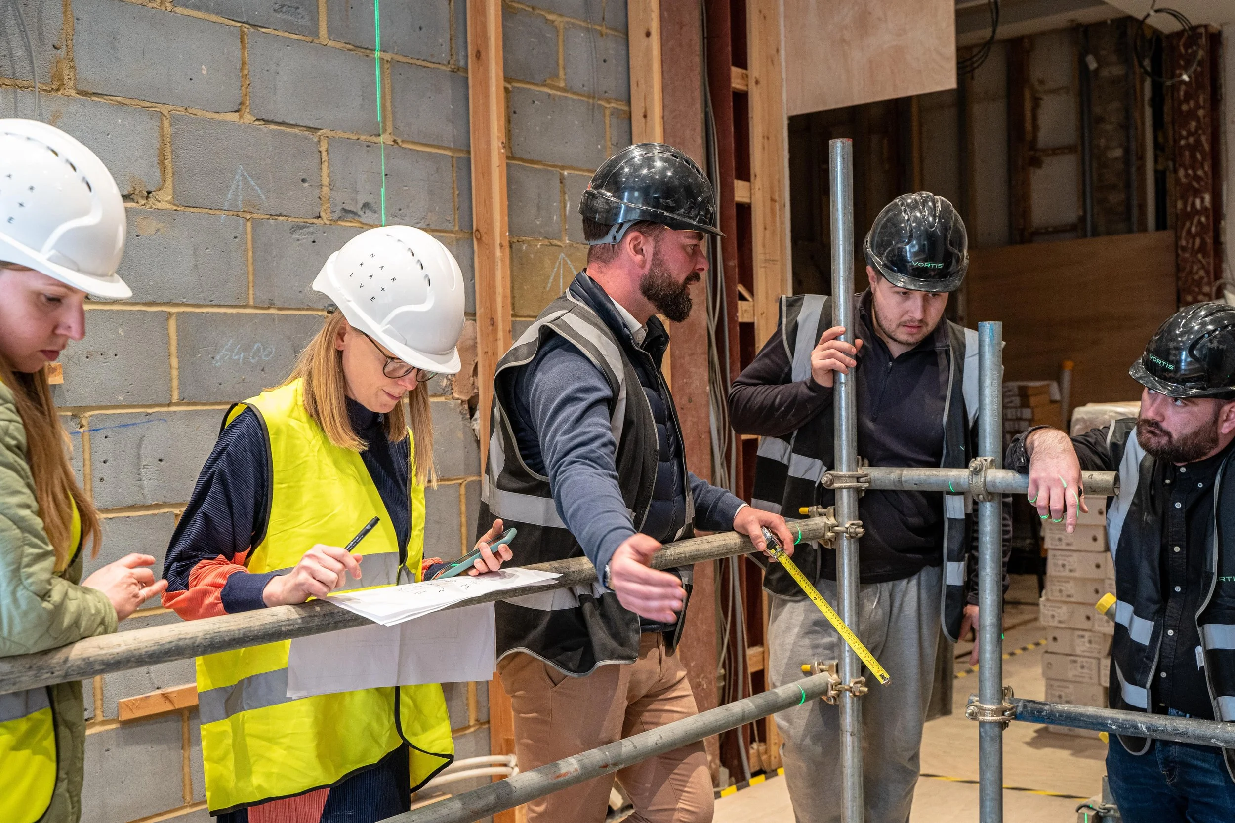 Group of construction workers and engineers wearing safety helmets and high-visibility vests working together at a construction site indoors. They are gathered around scaffolding, looking at plans and using measuring tools.