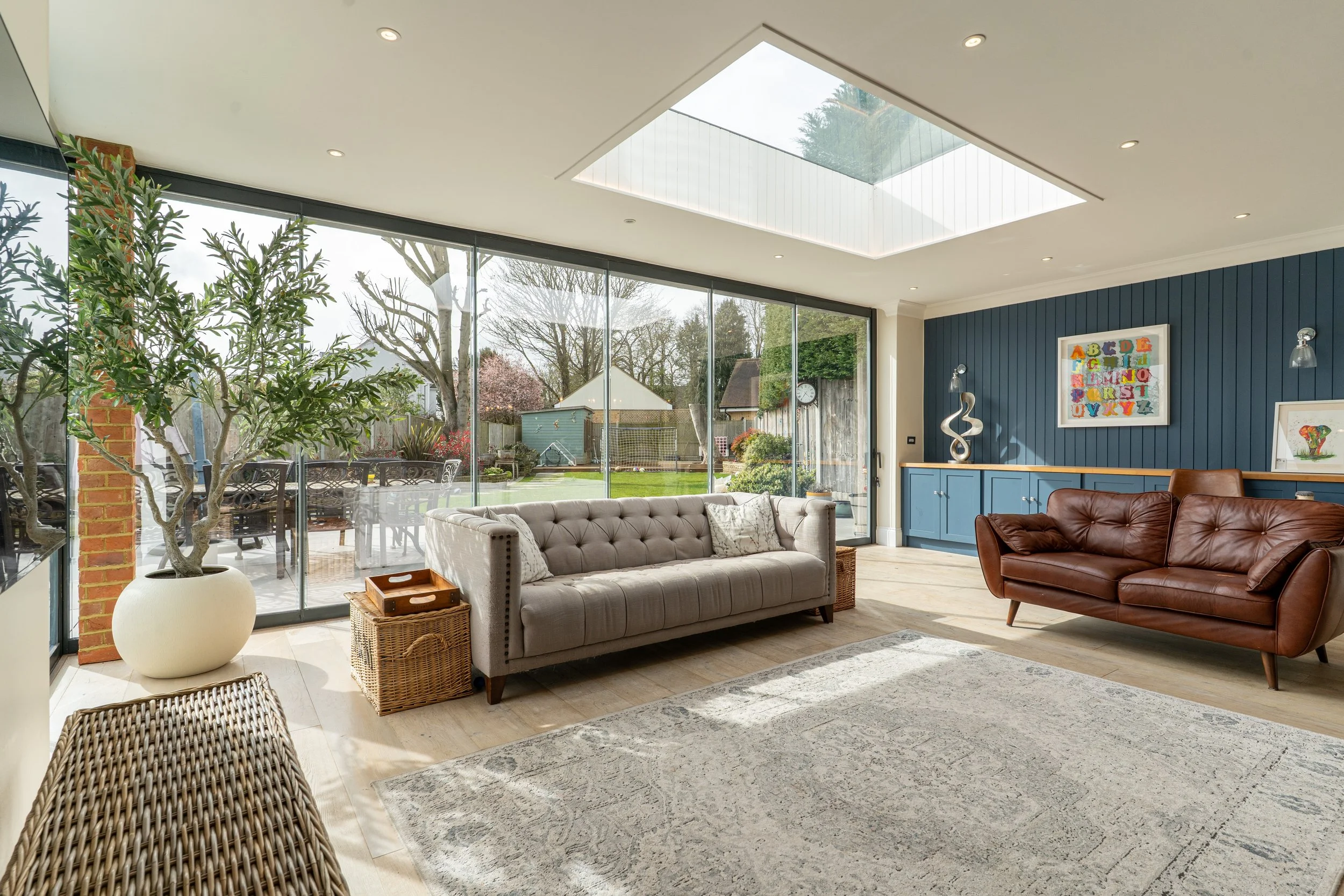Living room with large glass windows, three sofas, a wooden coffee table, a decorative sculpture, colorful framed artwork, and a view of a backyard with trees and outdoor furniture.