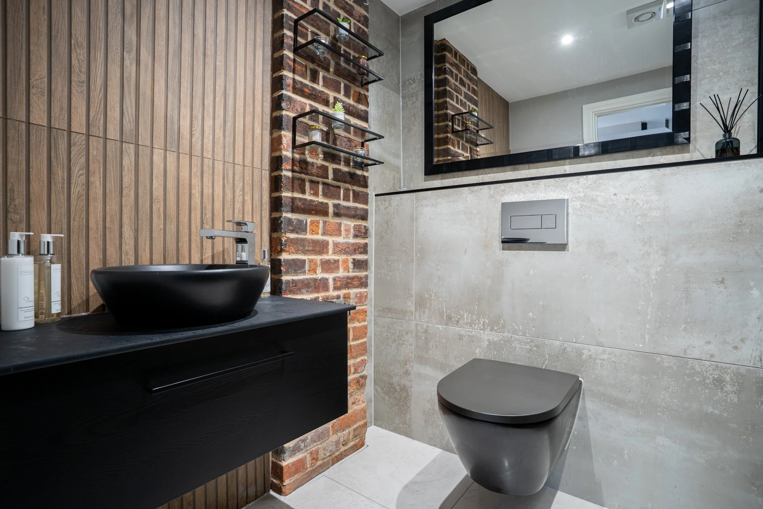 Modern bathroom with a black sink and a wooden vanity, exposed brick wall, large mirror, toilet, and decorative shelves with small plants, bottles, and reed diffuser.