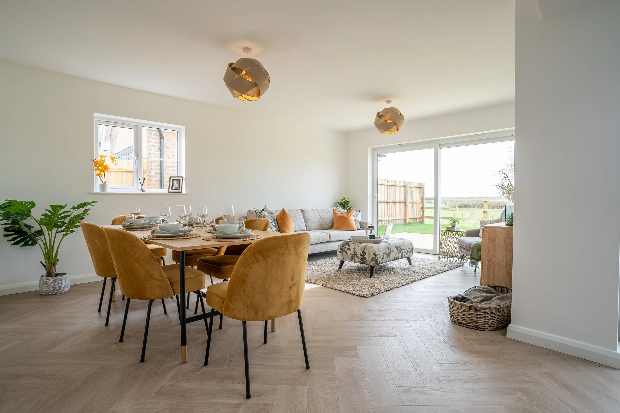 Open-concept living and dining room with large windows and sliding glass door, hardwood flooring, mustard velvet dining chairs, round dining table set with white dishes, glassware, living area with sofa, throw pillows, ottoman, plants, and a view of 