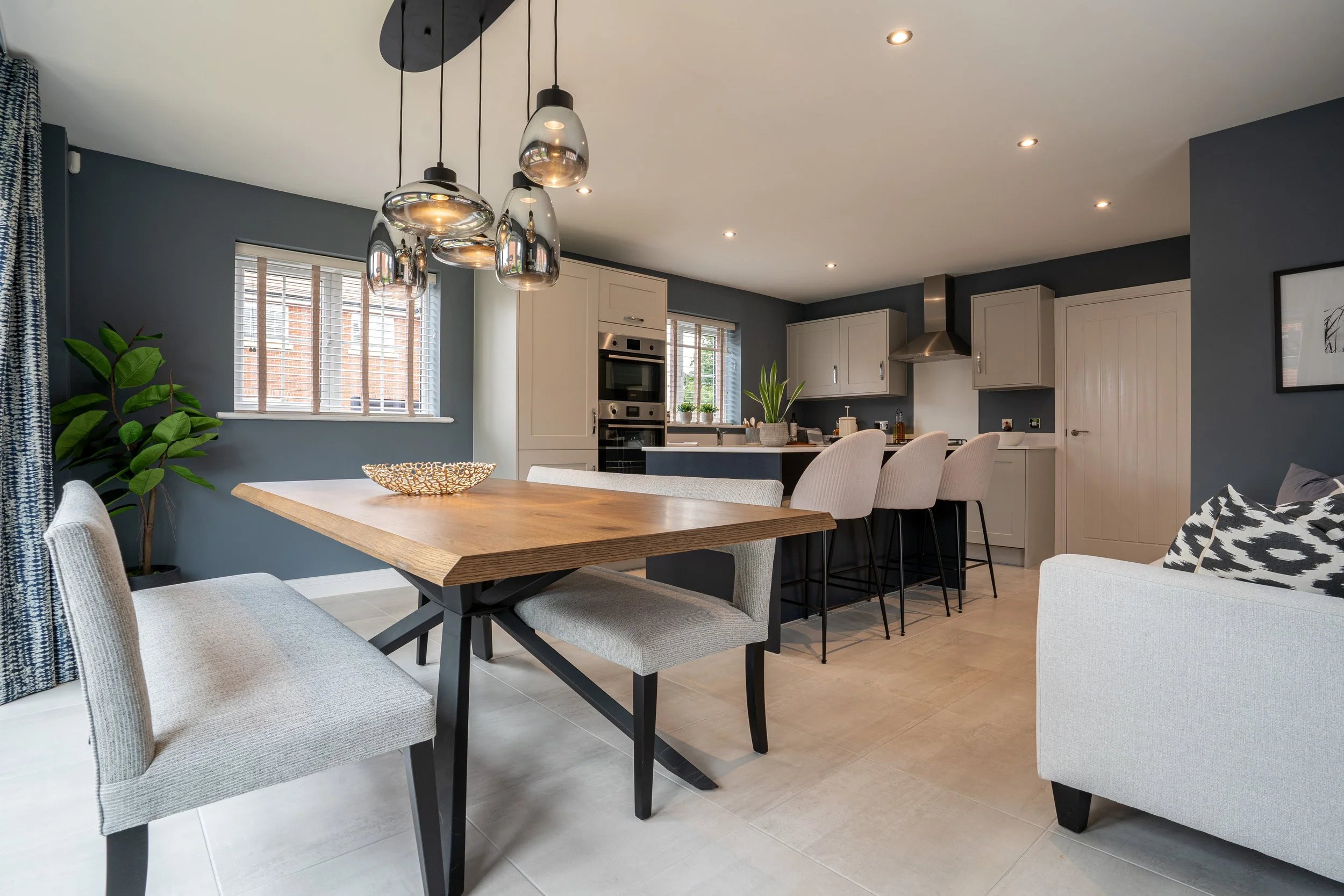 Modern kitchen and dining area with a wooden table, light beige upholstered chairs, gray walls, white cabinets, stainless steel appliances, a large window with blinds, green plants, and pendant lights.