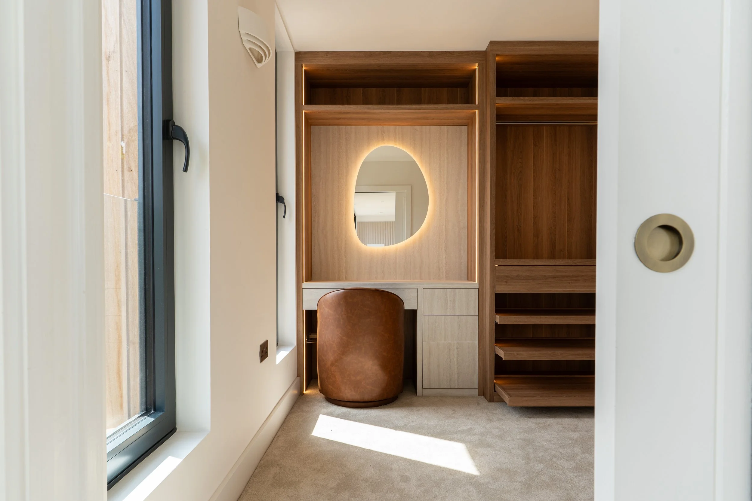 A modern walk-in closet with wooden shelves, a mirror with lighting, a leather ottoman, a window, and a door.