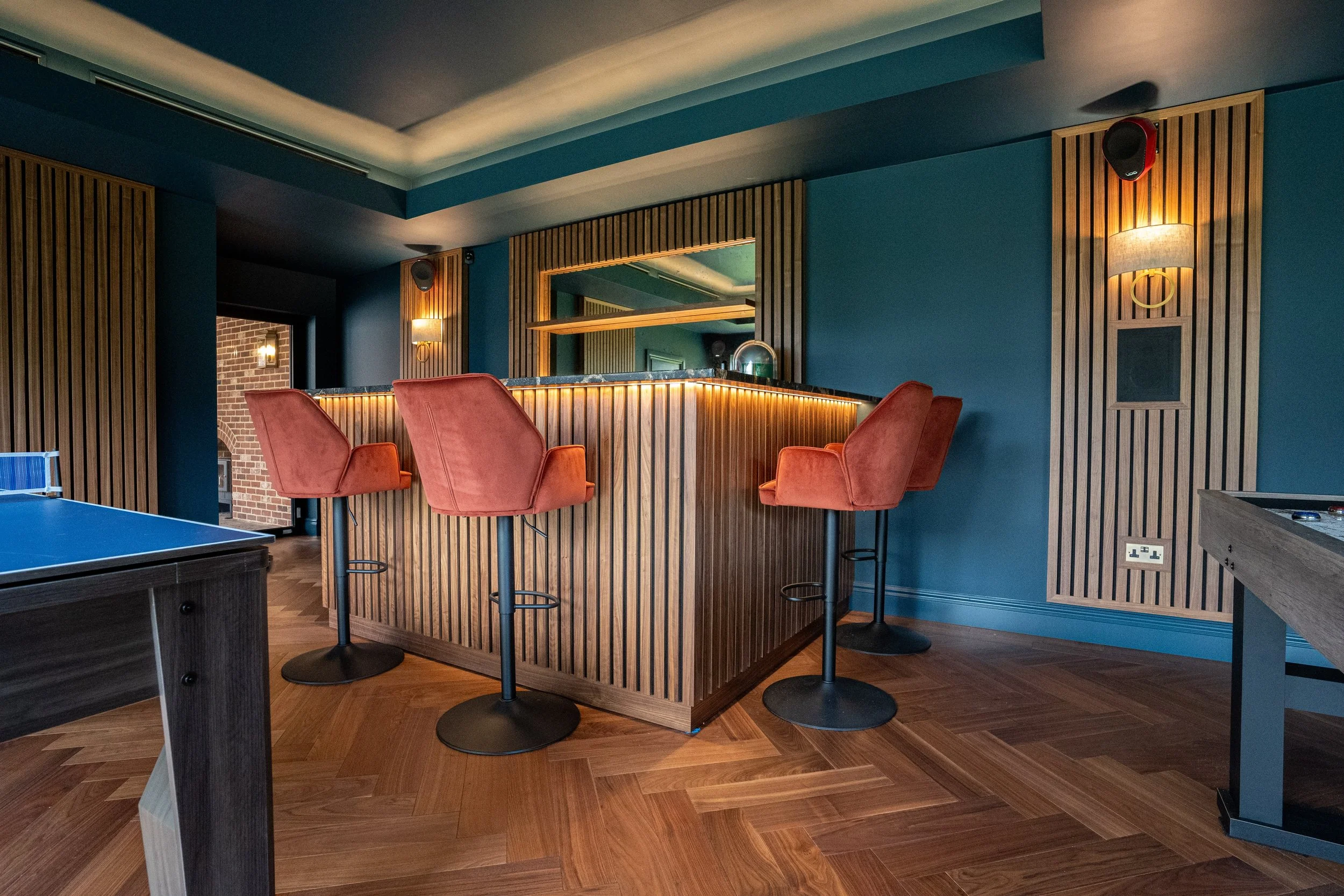 Interior of a bar or lounge with a wooden counter, four red barstools, and blue walls with wooden slat accents and wall sconces.