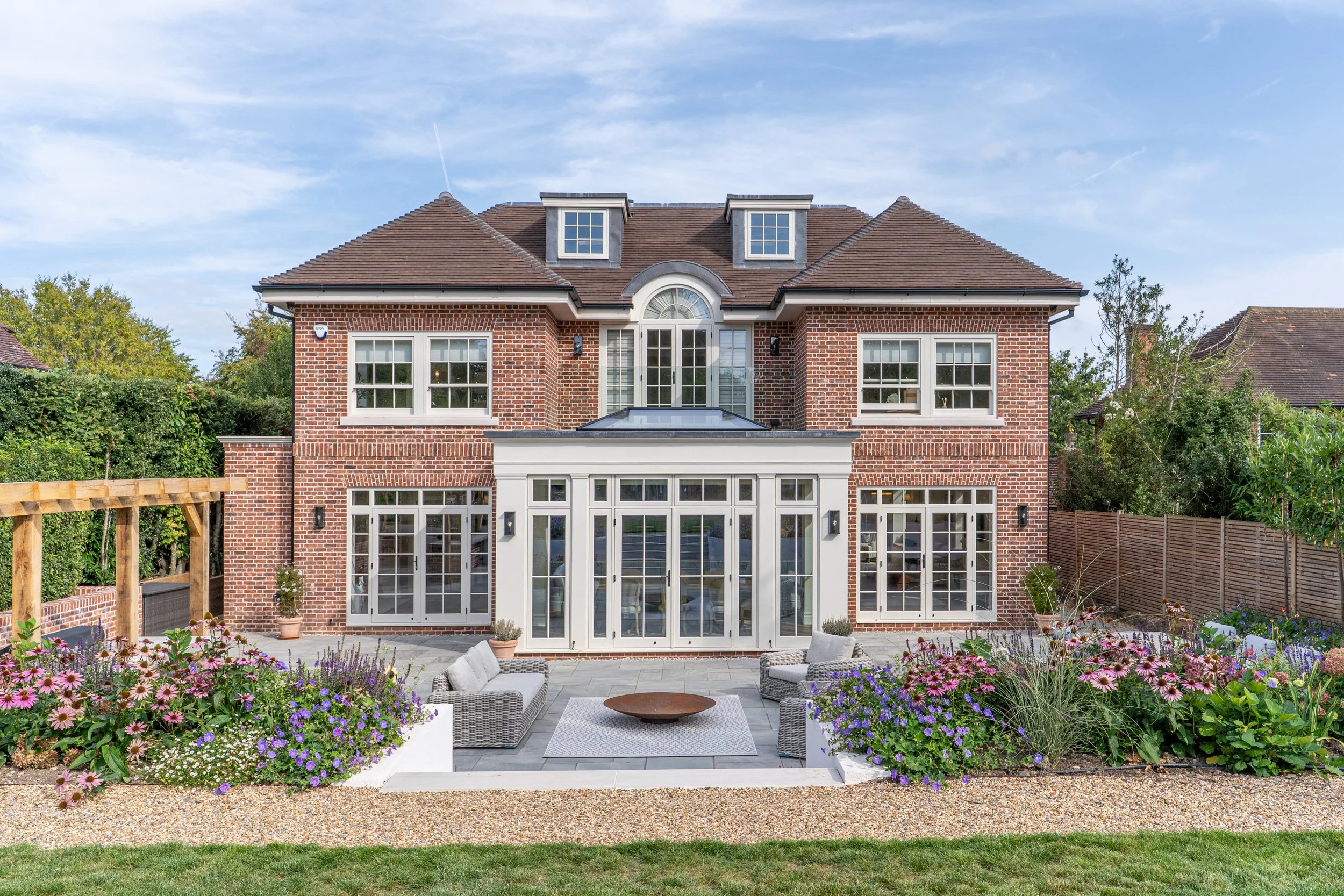 Backyard patio of a brick house with large windows, surrounded by flower beds, outdoor furniture, and a gravel walkway.