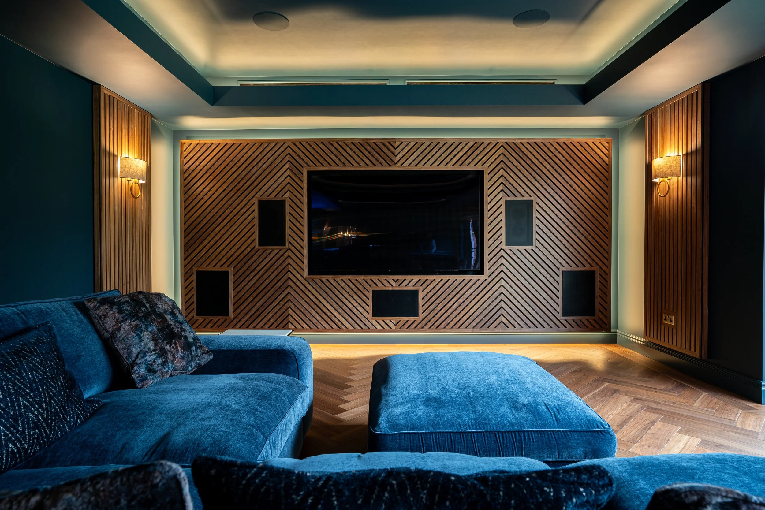 A living room with blue velvet sofas, some with patterned cushions. A wooden paneled wall with a mounted flat-screen TV and black acoustic panels. Two wall-mounted lamps are on either side, with warm lighting. The ceiling has recessed lighting and tw