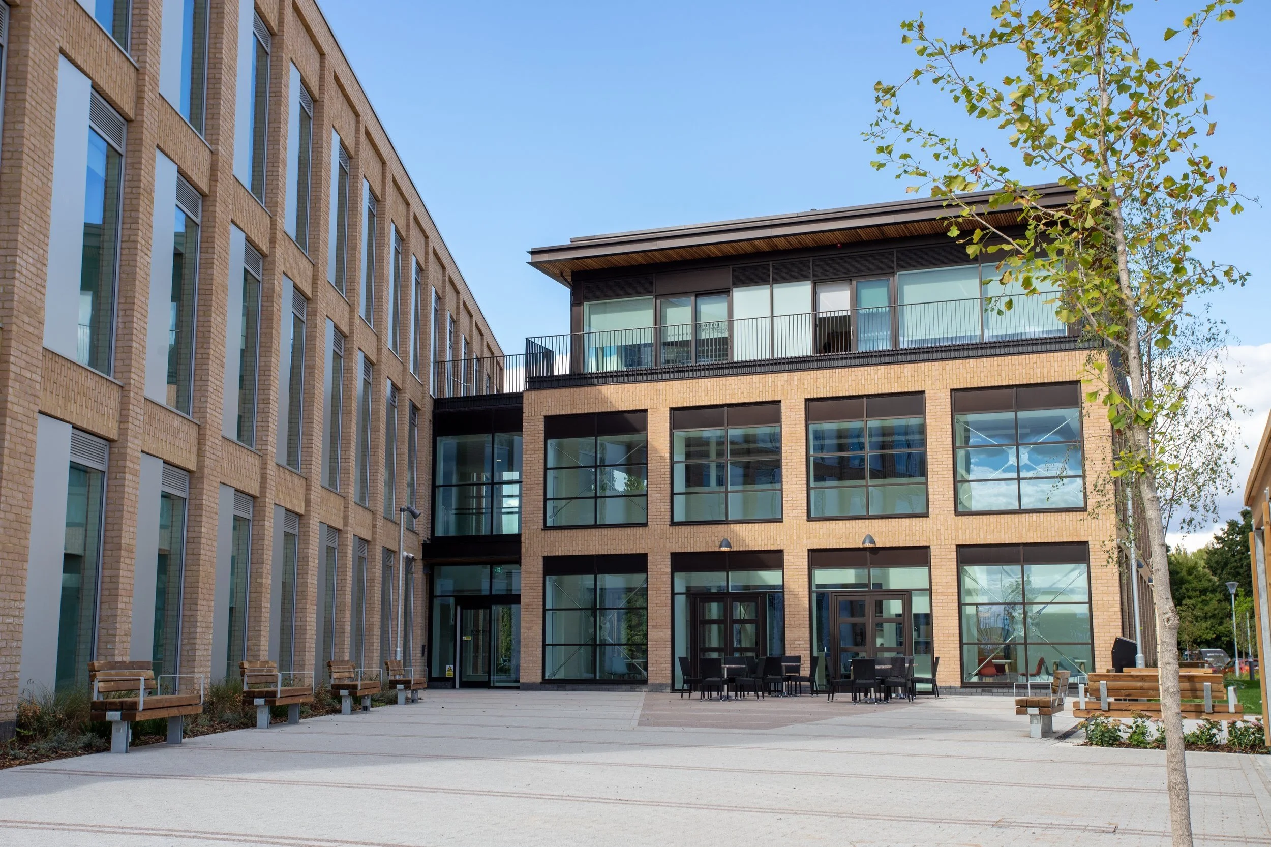 Modern brick building with large glass windows and a small outdoor seating area with benches and chairs, trees, and a clear blue sky