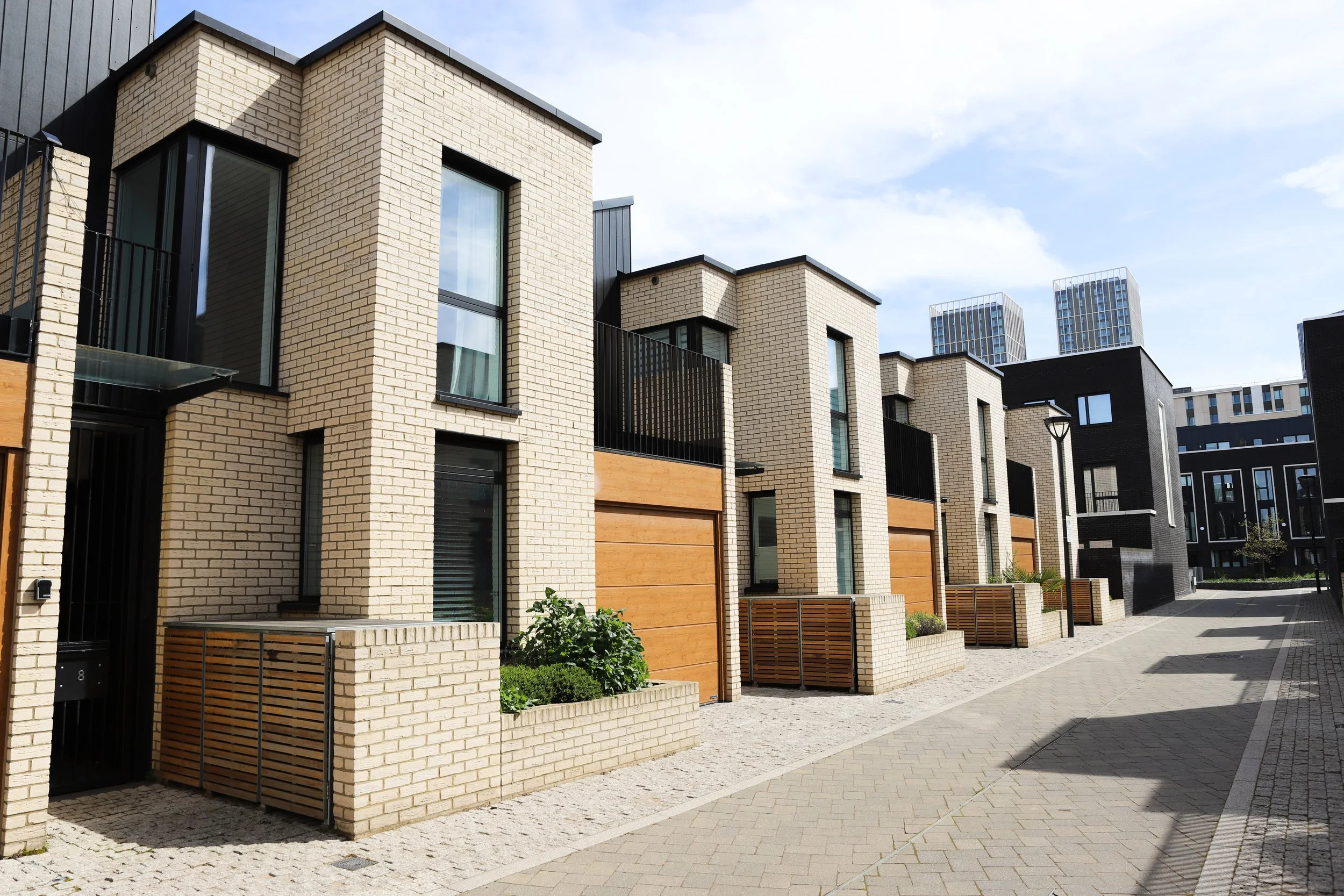 Modern residential buildings with brick and wood panels, large windows, and small balcony fences, on a paved street under a partly cloudy sky.