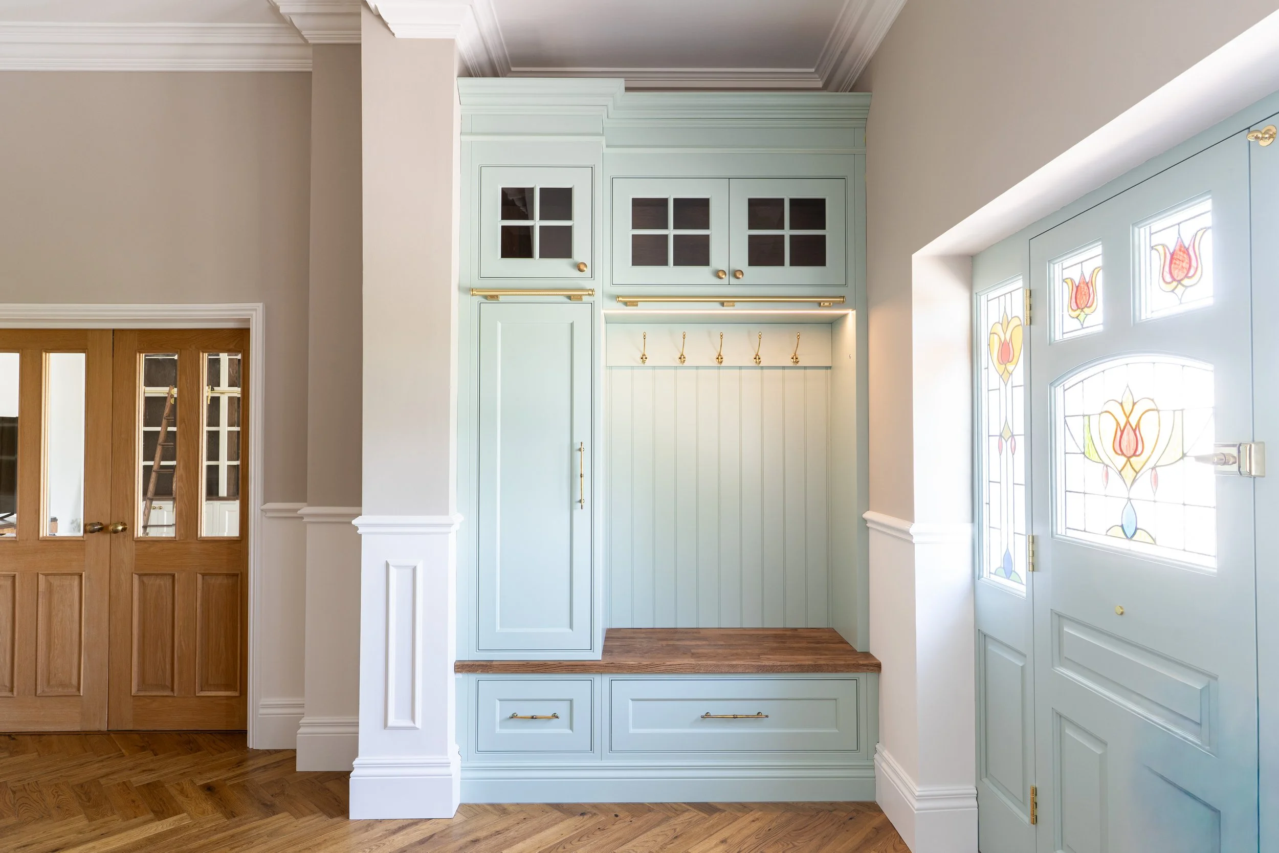 Entryway with built-in light teal cabinetry, gold hardware, and stained glass front door, adjacent to wooden double doors with glass panes.