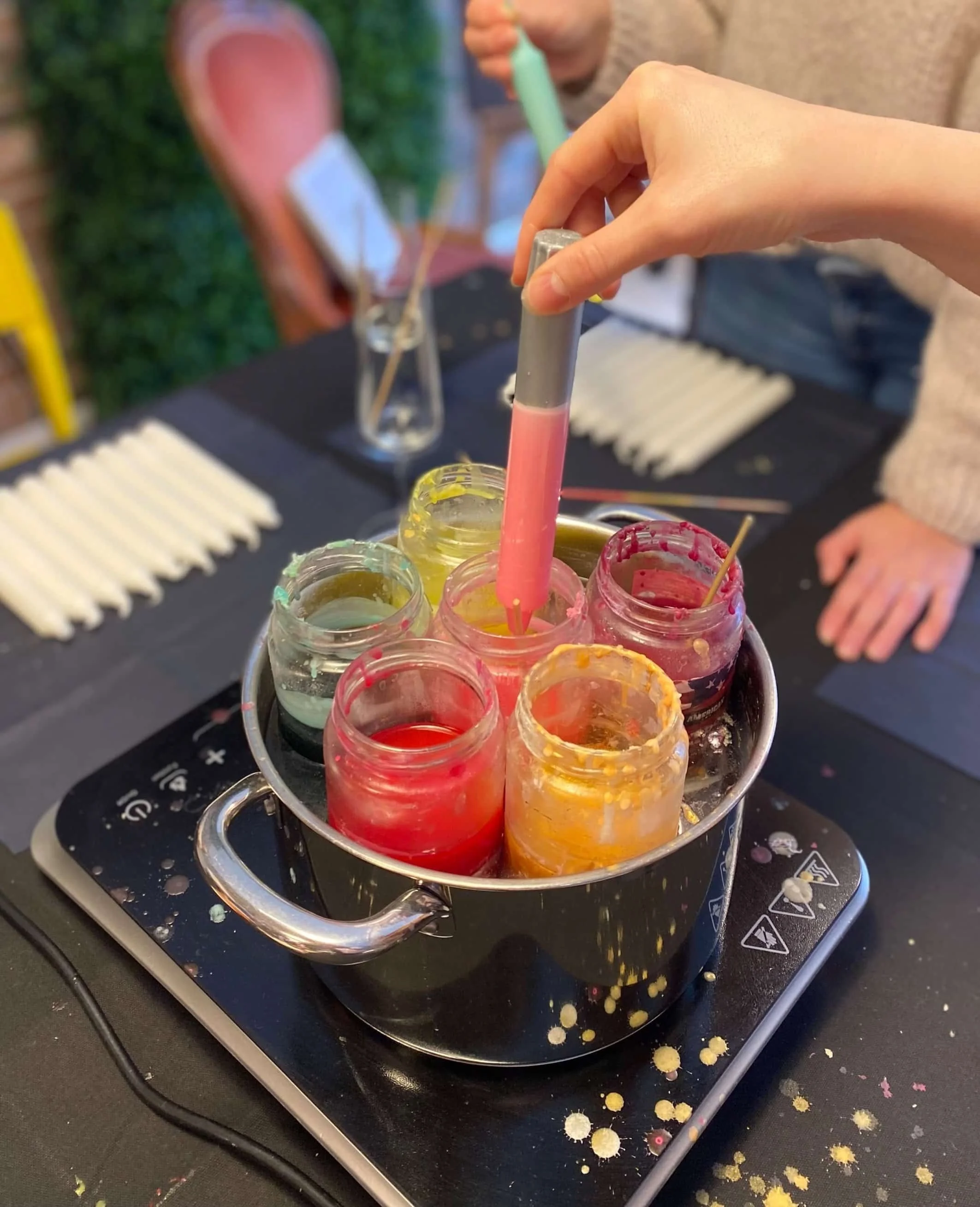 Person, die farbigen flüssigen Wachs in kleine Gläser mit Pipette füllt, auf einem schwarzen Tisch, im Hintergrund eine Person und eine Vase mit einer Blume.