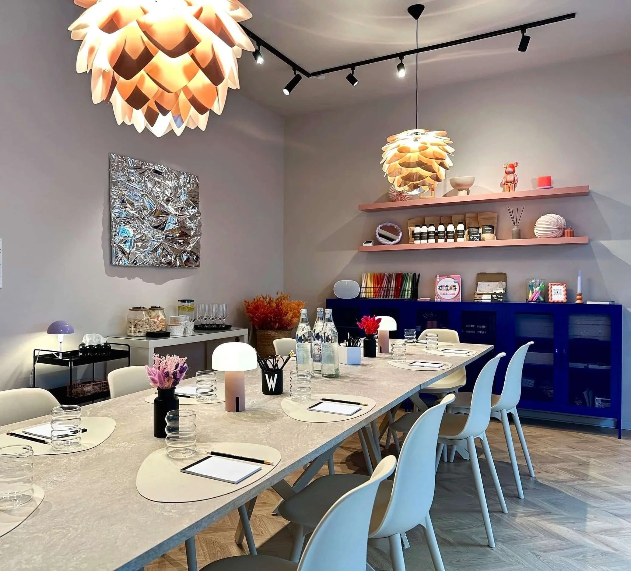 Lange Tischtafel mit Notizbücher, Bleistiften, Lampen, Wasserflaschen und Blumenvasen drauf, im Hintergrund blauer Schrank, Regalböden mit Deko