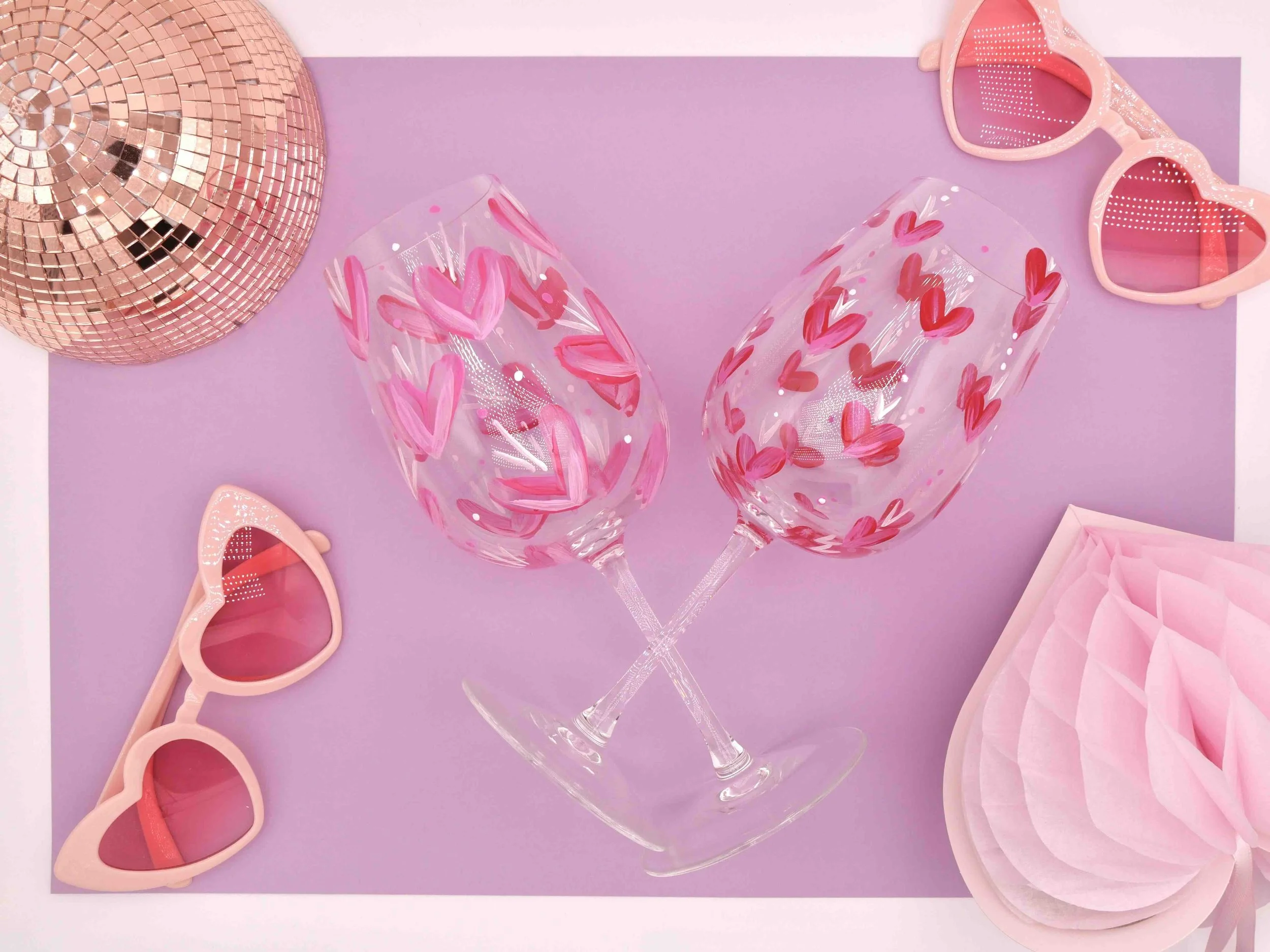 Zwei Weingläser bemalt mit pinken Herzen in verschiedenen Größen daneben zwei rosa Brillen in Herzform, rosa Diskokugel und Wabenball in Form eines Herzens