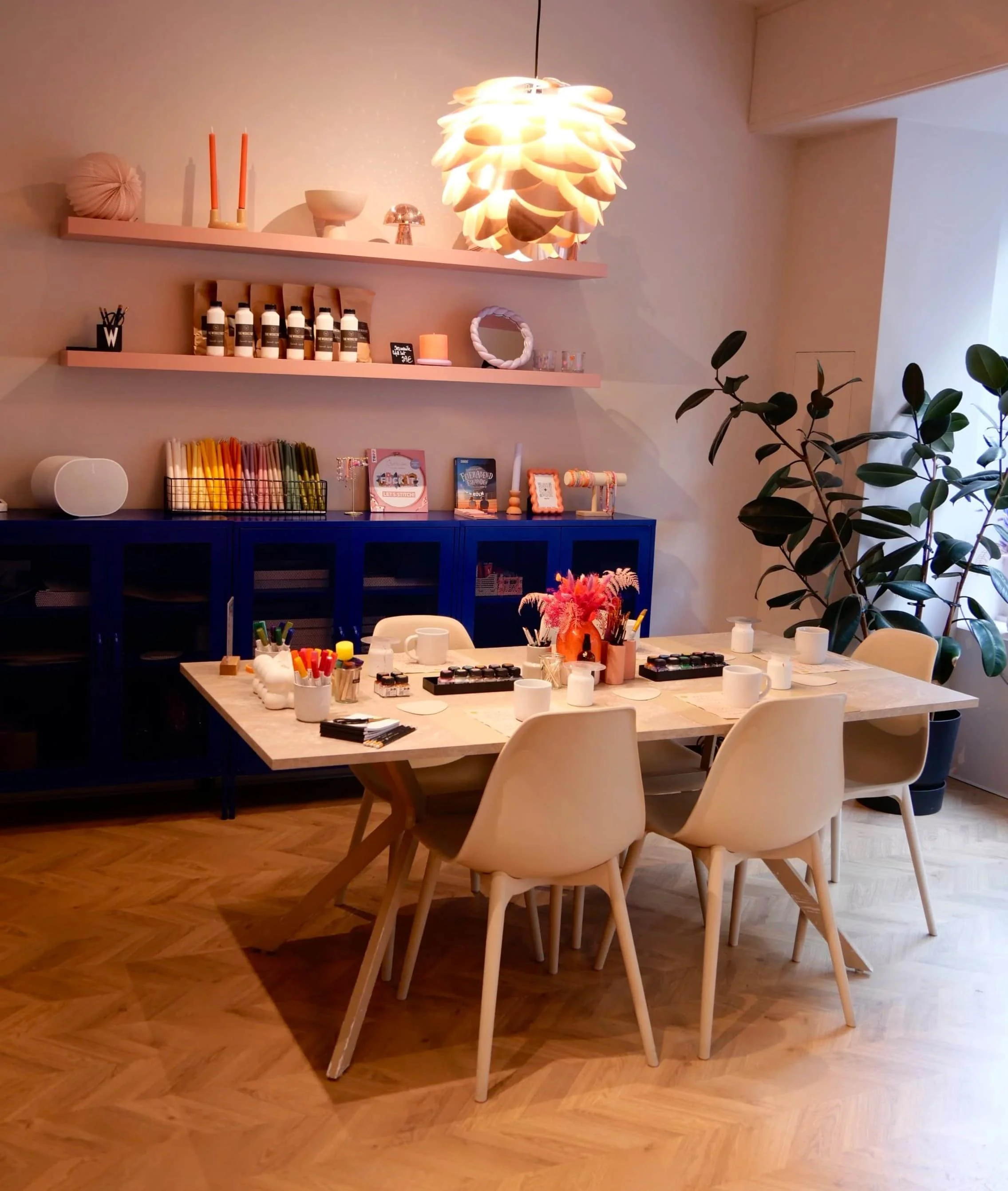 Kreativraum mit einem Tisch mit Malutensilien und fünf Stühlen vor einem blauen Regal mit Dekoration und Kerzen