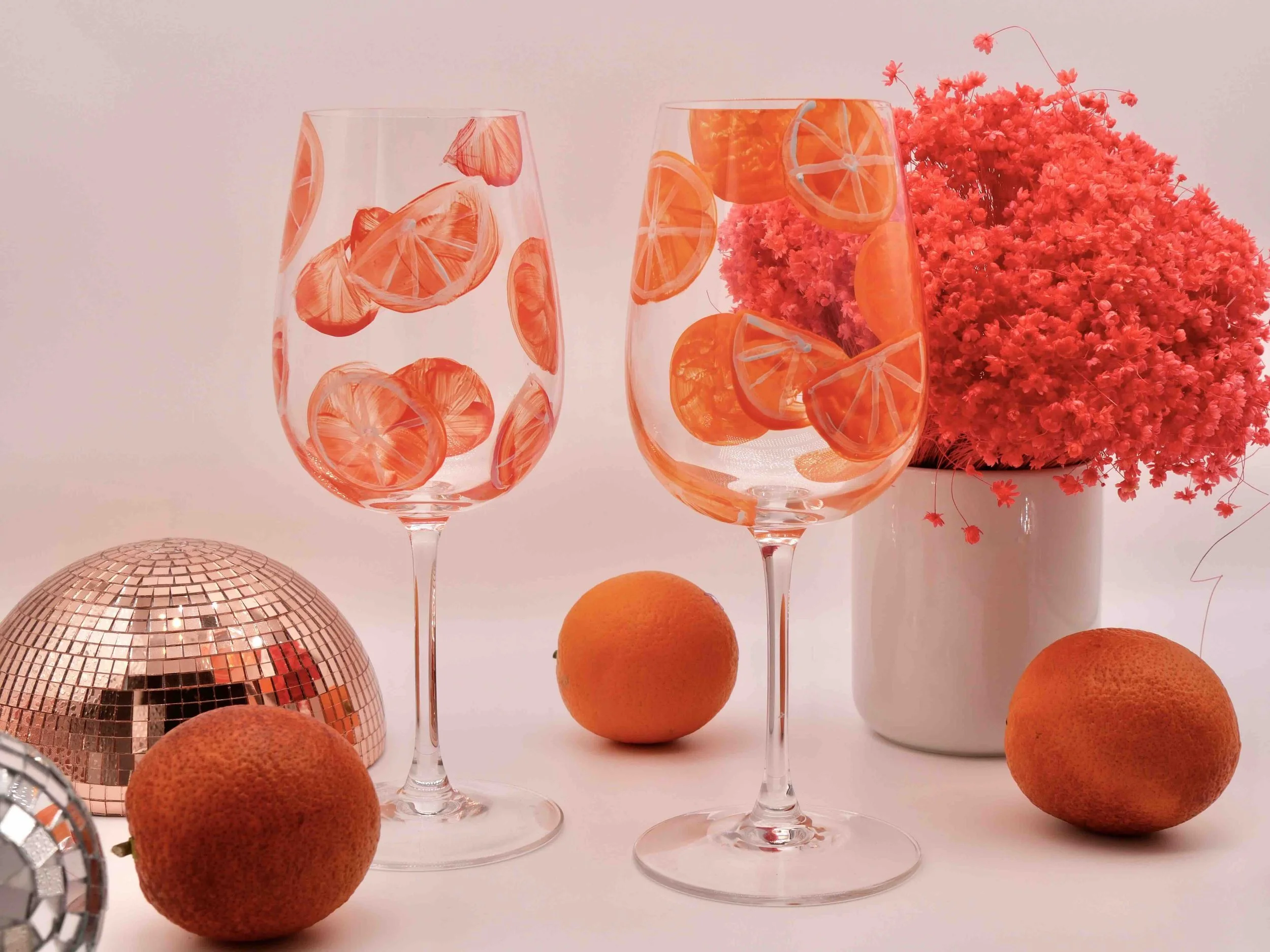 Zwei mit Orangenmotiven bemalte Weingläser, Orangen und orangene Blumen sowie Diskokugel im Hintergrund