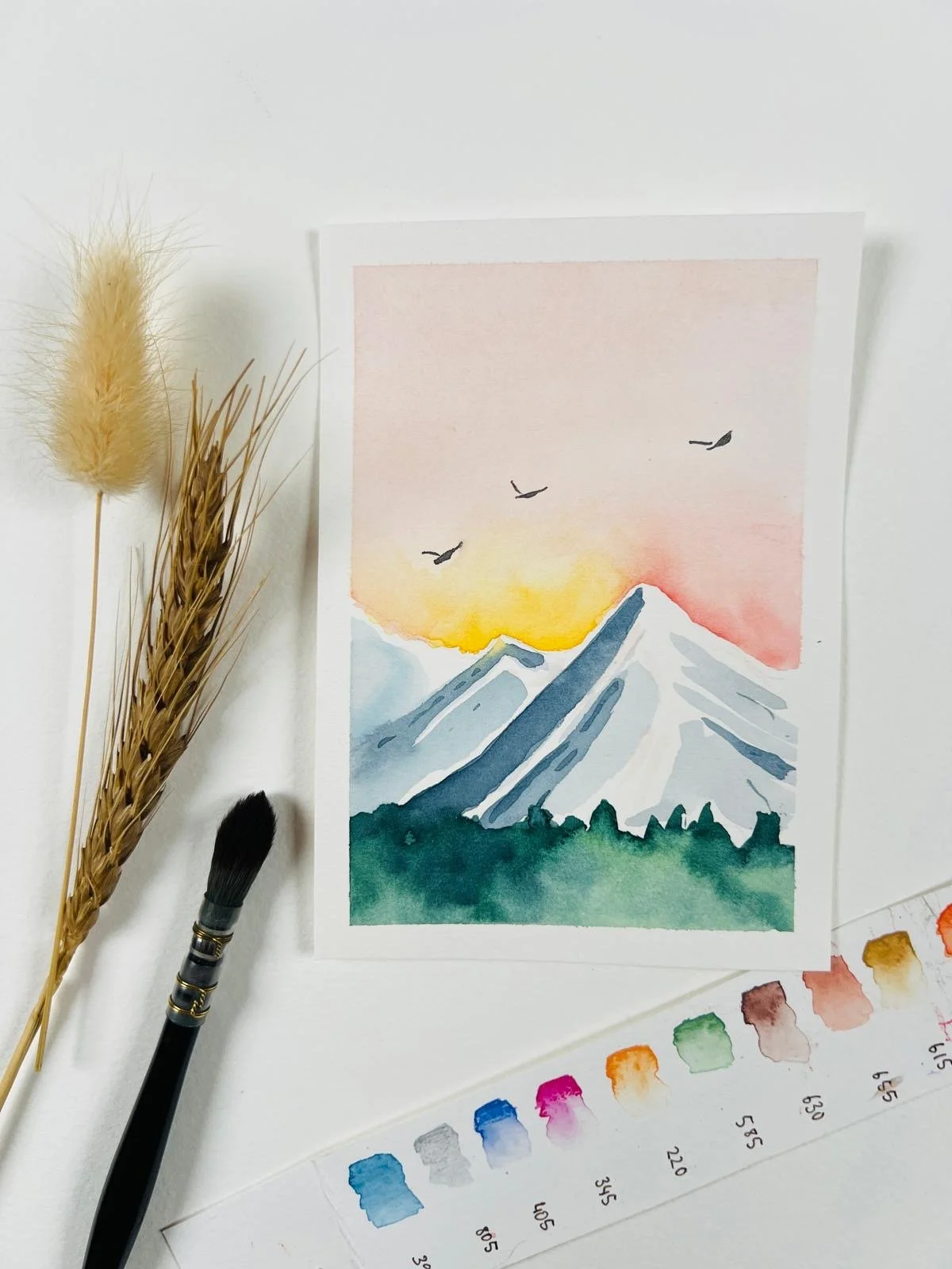 Ein Aquarellgemälde von schneebedeckten Bergen im Sonnenaufgang mit drei fliegenden Vögeln am Himmel, platziert auf einem weißen Hintergrund – daneben liegen getrocknete Gräser, ein schwarzer Pinsel und eine Farbpalette.