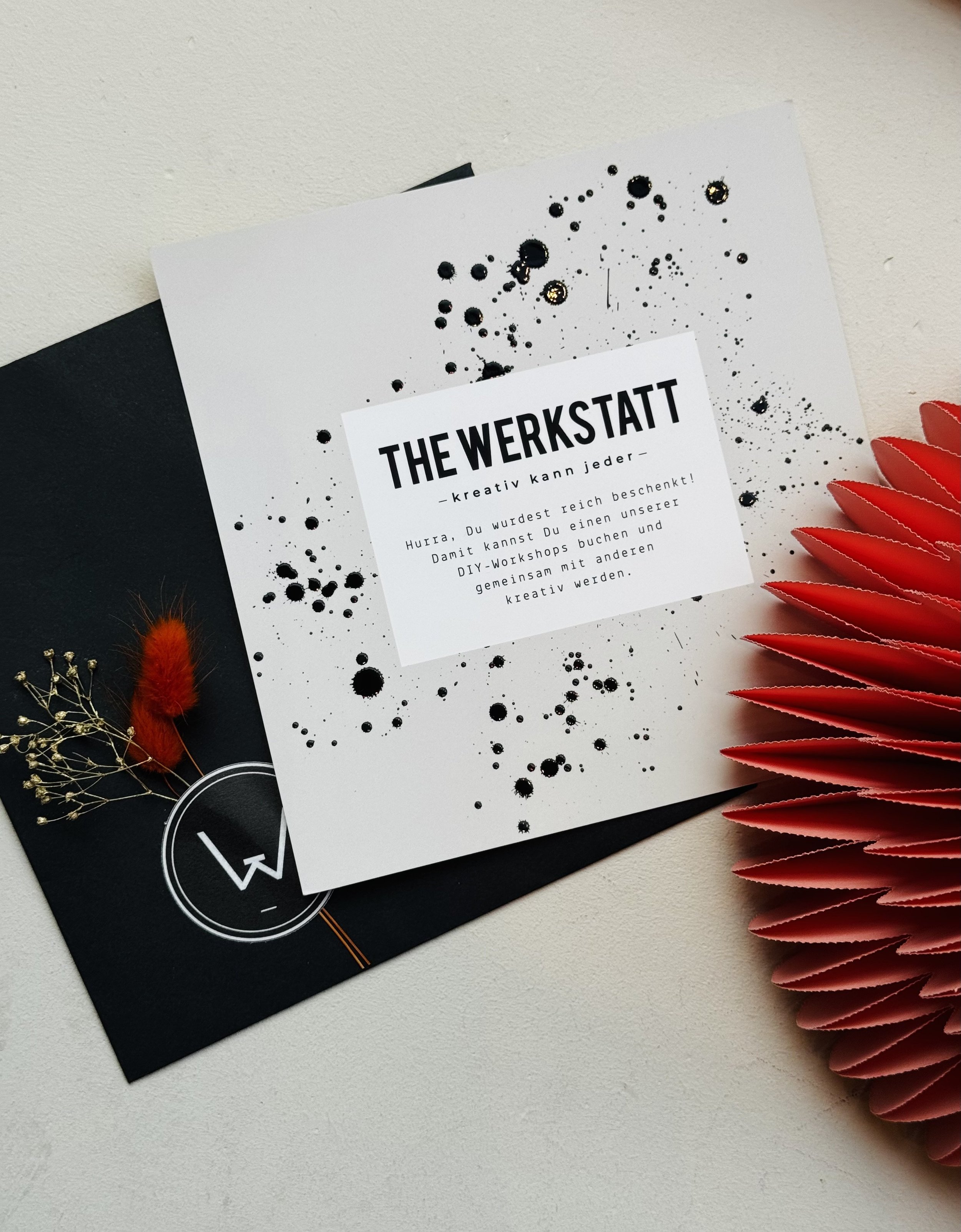 Ein Gutschein mit der Aufschrift 'THE WERKSTATT - Kreativ kann jeder', umgeben von schwarzen und goldenen Akzenten, liegt auf einem schwarzen Umschlag, neben einer roten Papierdekoration und getrockneten Blumen.